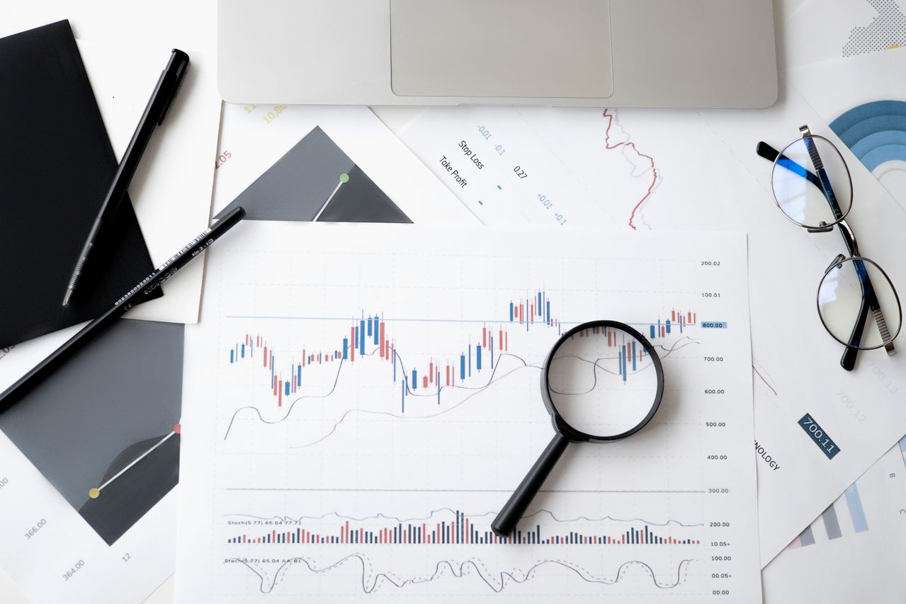 Tampilan atas meja kerja berisi grafik analisis saham dan keuangan, kaca pembesar, laptop, serta alat tulis untuk pemantauan bisnis