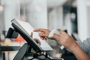 aplikasi-kasir-simon-kadula-pos-touchscreen-kasair Kasir menekan layar sentuh mesin POS dan memegang struk pembayaran belanja menggunakan Kasair POS
