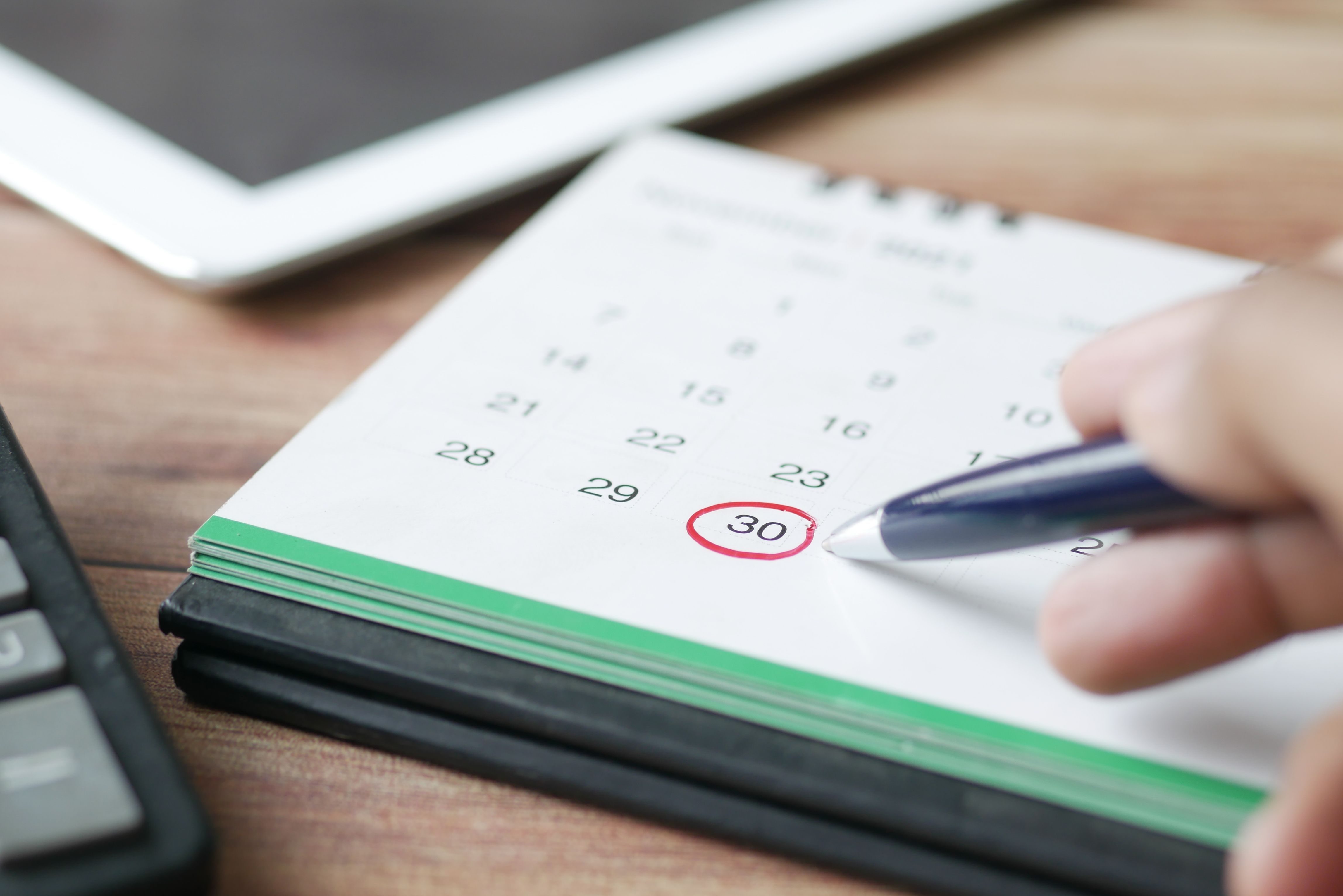Tangan memegang pena melingkari tanggal 30 pada kalender meja di atas meja kayu