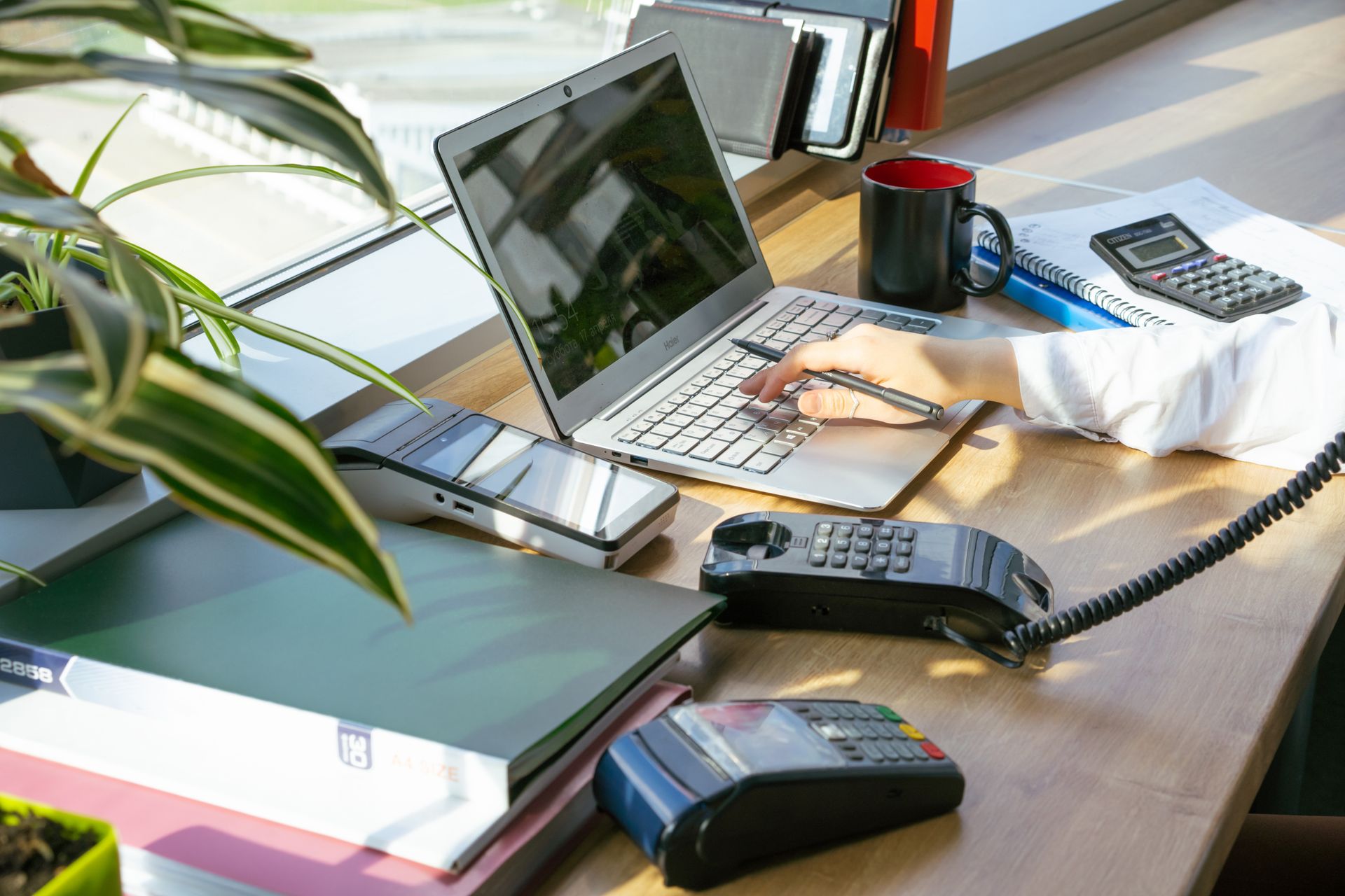 Seseorang sedang mengelola transaksi dan keuangan bisnis menggunakan laptop serta mesin EDC pembayaran di meja kerja