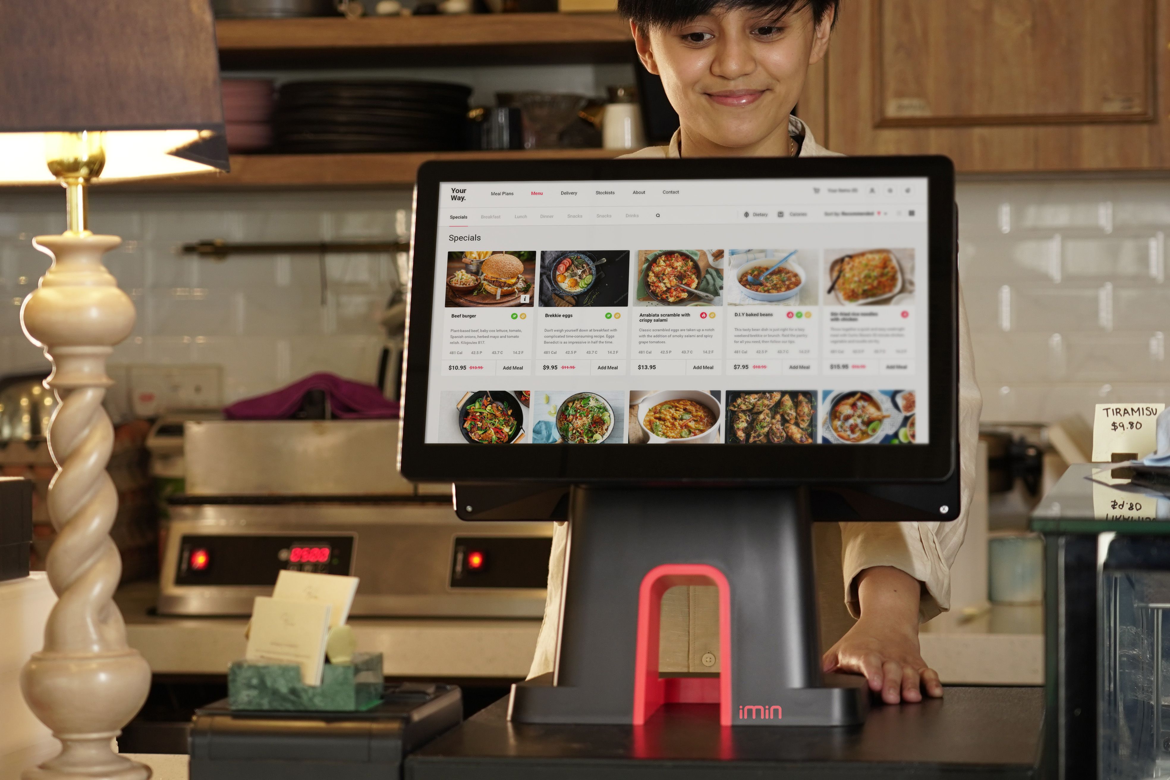 Staf kasir restoran tersenyum di balik layar monitor POS digital yang menampilkan menu makanan.