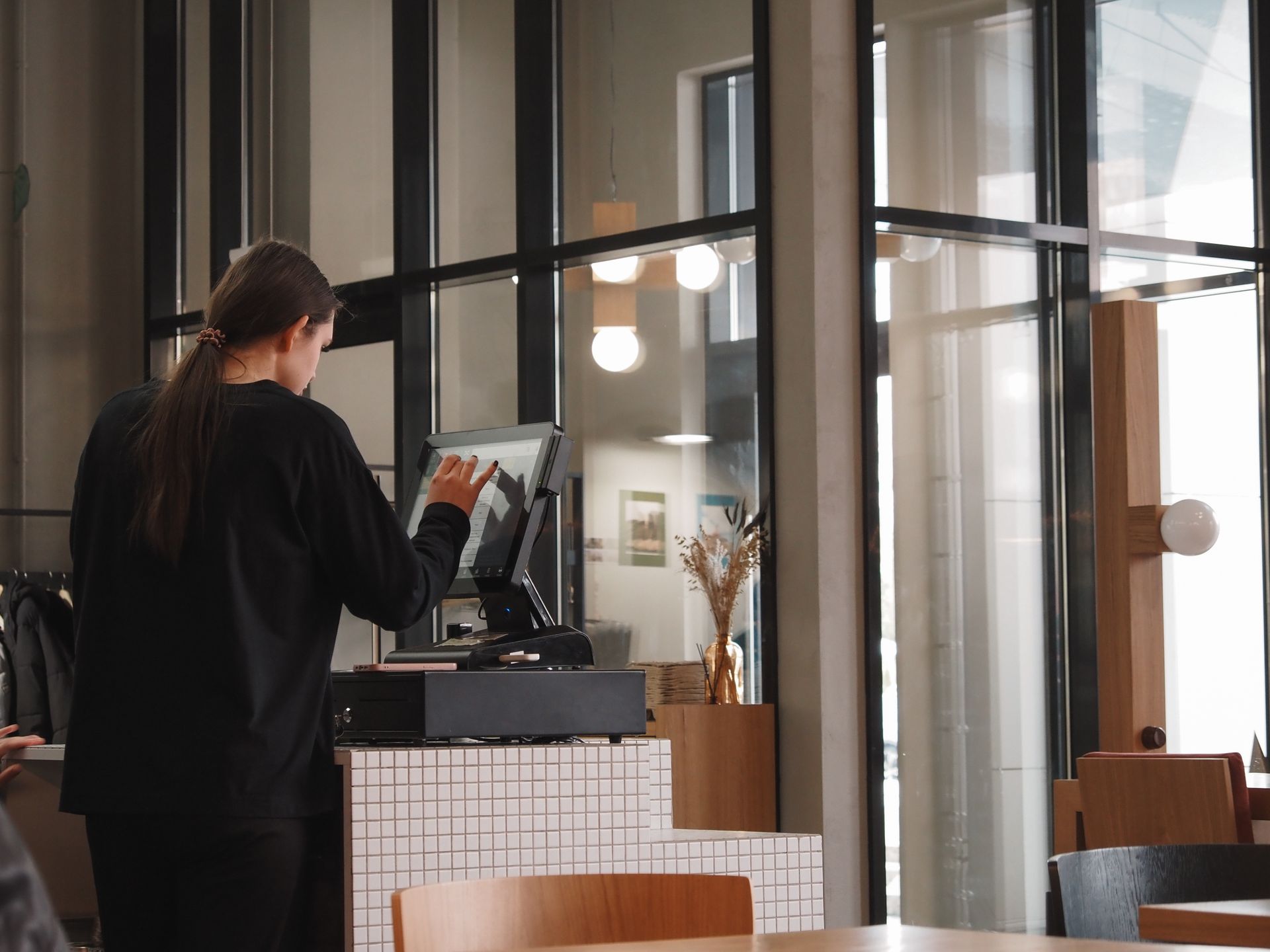 Kasir perempuan sedang menekan layar sentuh terminal mesin POS di meja kasir kafe modern