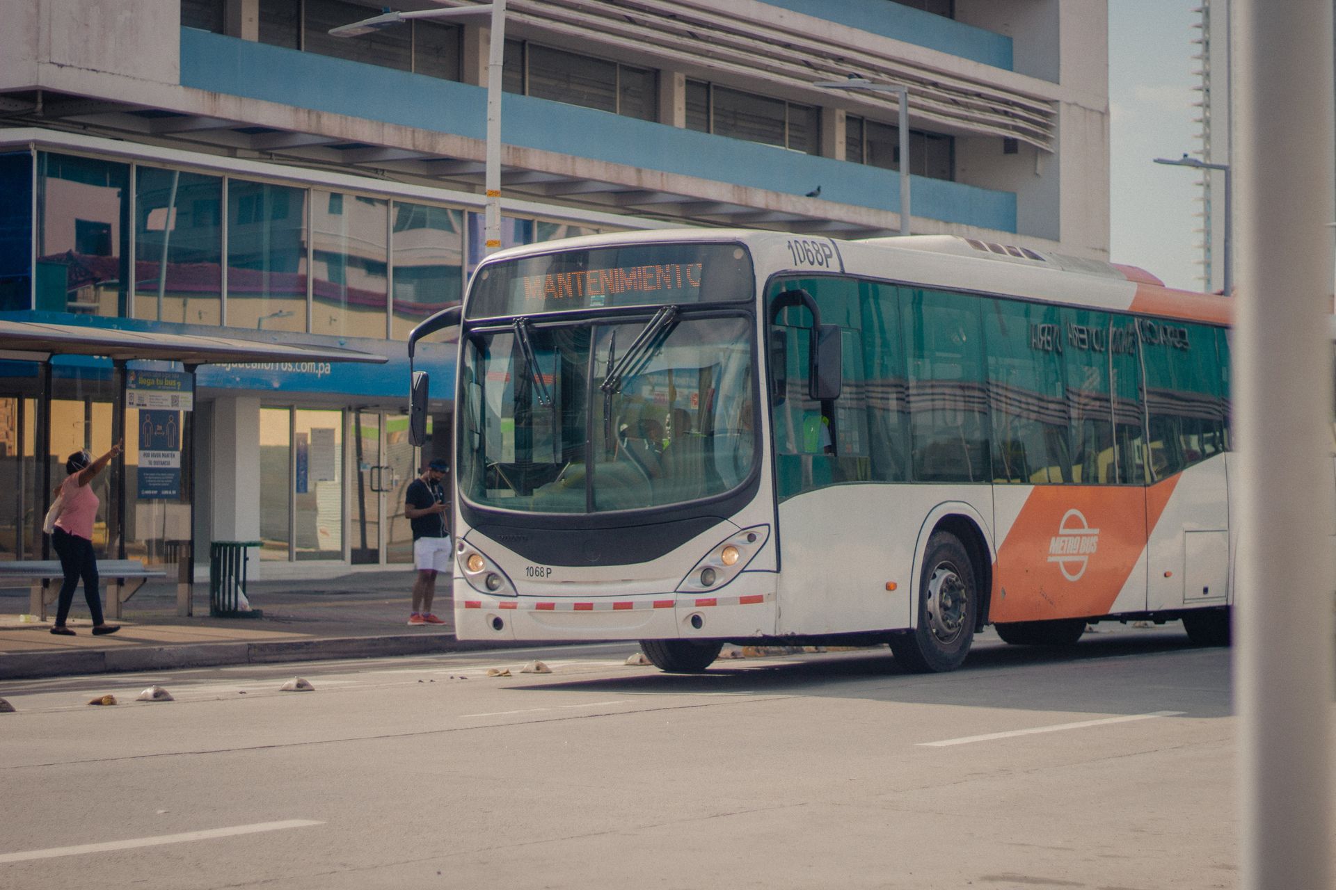 Bus kota berwarna putih dan oranye sedang melintas di depan halte bus transportasi publik