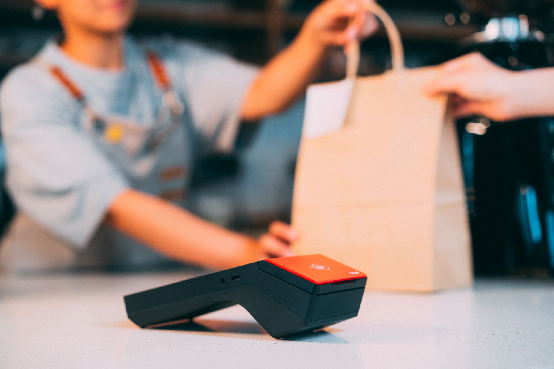 Perangkat kasir portabel mobile POS di meja kasir saat staf menyerahkan kantong pesanan takeaway kepada pelanggan dengan Kasair POS