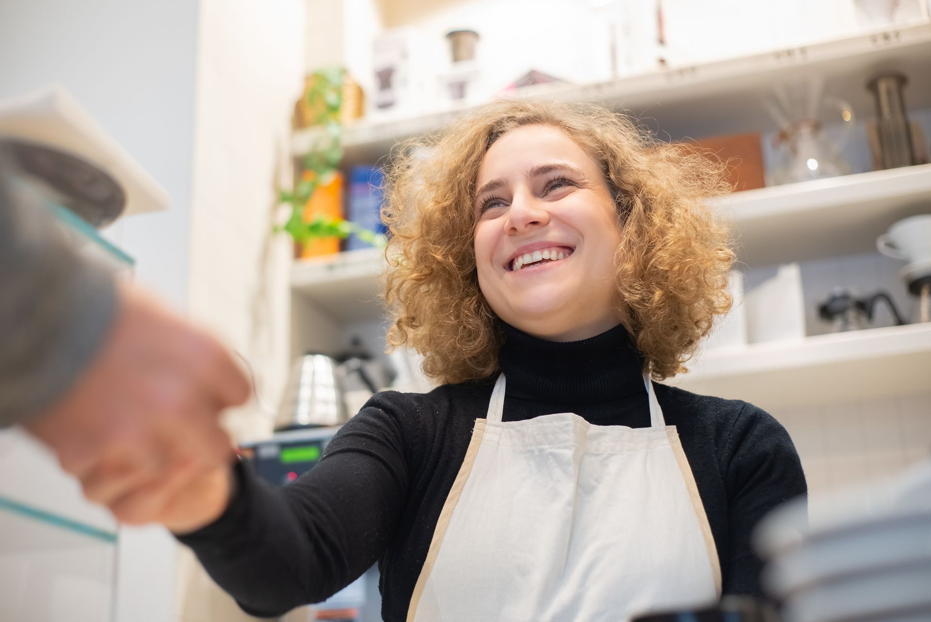 Barista wanita berapron tersenyum ramah saat berjabat tangan dengan pelanggan di meja kasir kedai kopi