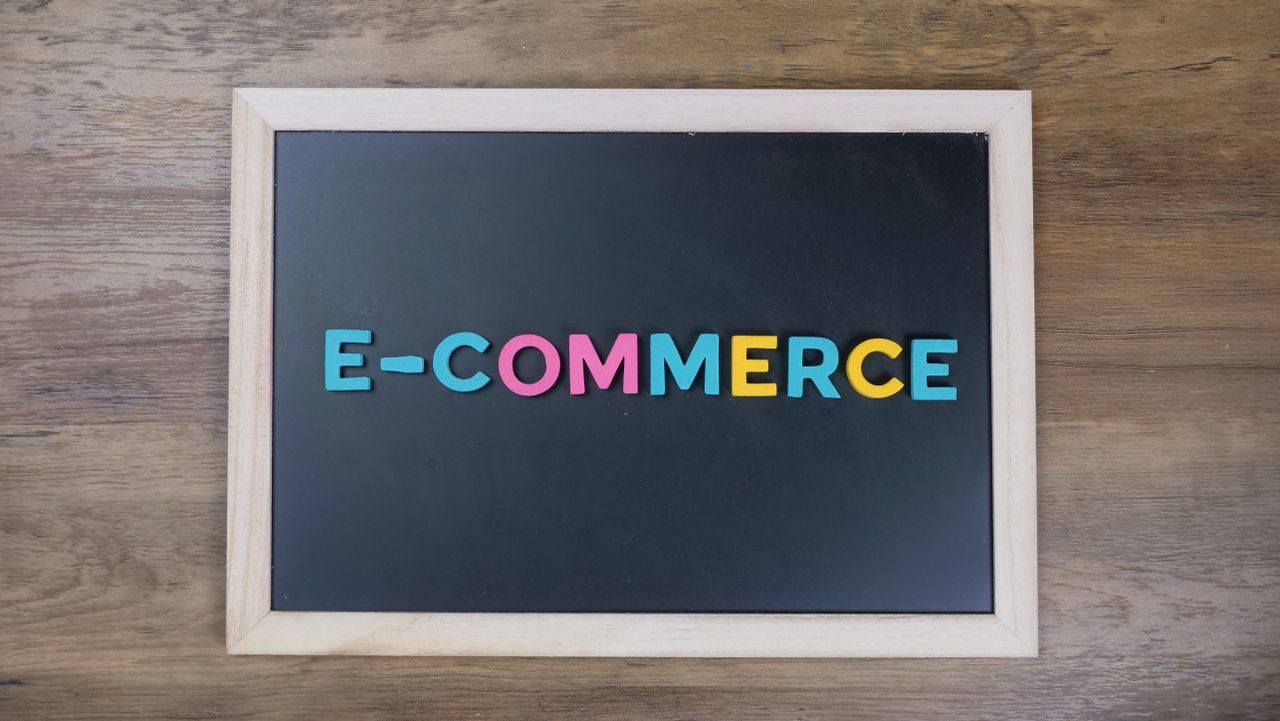 Papan tulis hitam berbingkai kayu dengan tulisan huruf warna-warni E-COMMERCE di atas meja kayu.