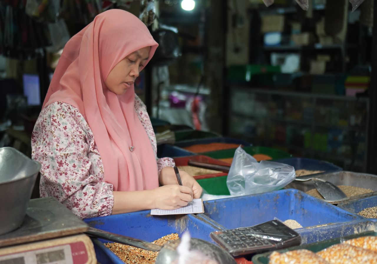 Wanita berhijab merah muda pedagang toko kelontong atau bahan pokok sedang mencatat transaksi di pasar tradisional.