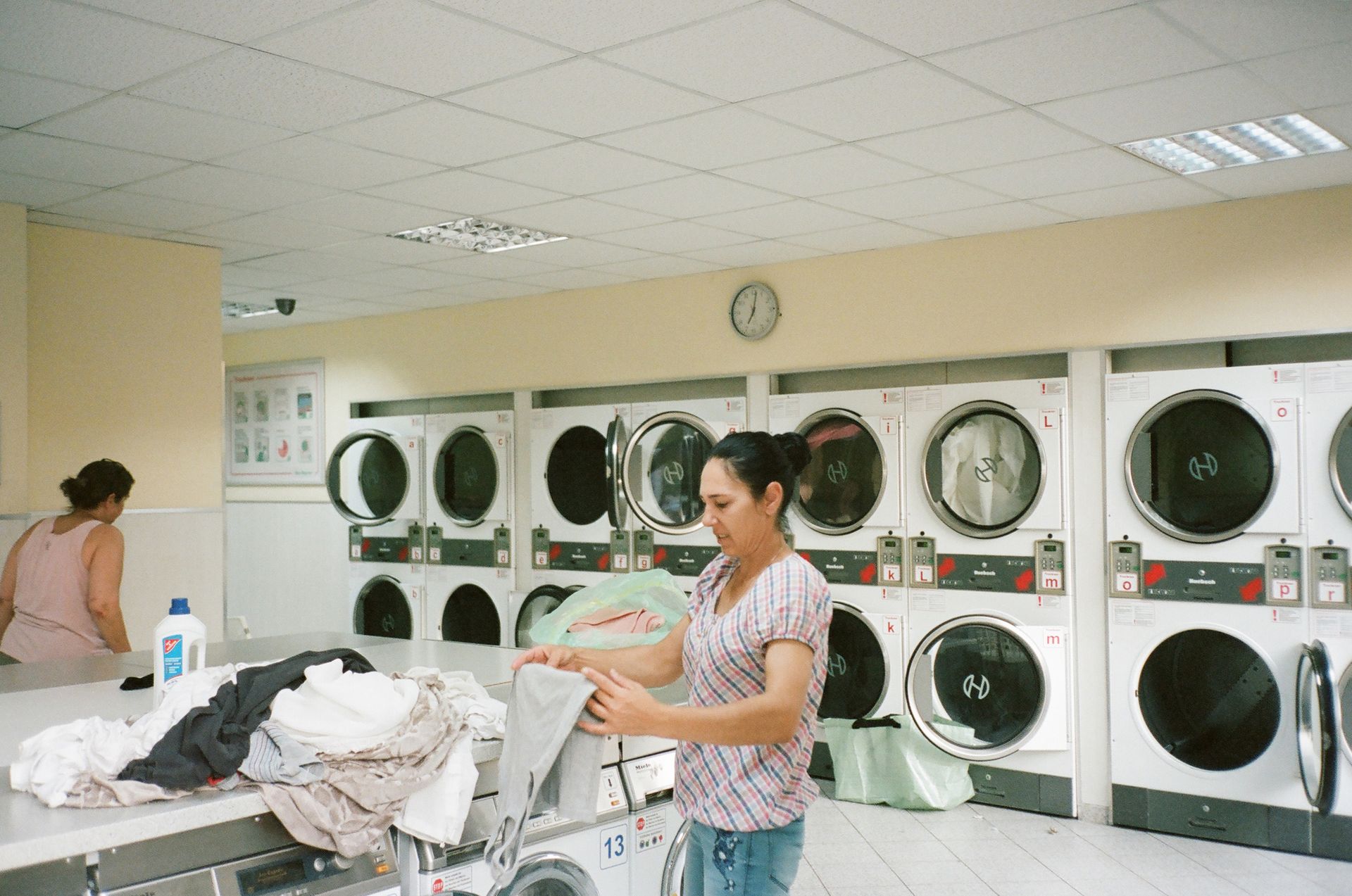 Aktivitas Layanan dan Operasional Usaha Laundry Wanita berkemeja kotak-kotak sedang melipat pakaian di atas meja dengan latar belakang deretan mesin cuci di ruangan usaha laundry