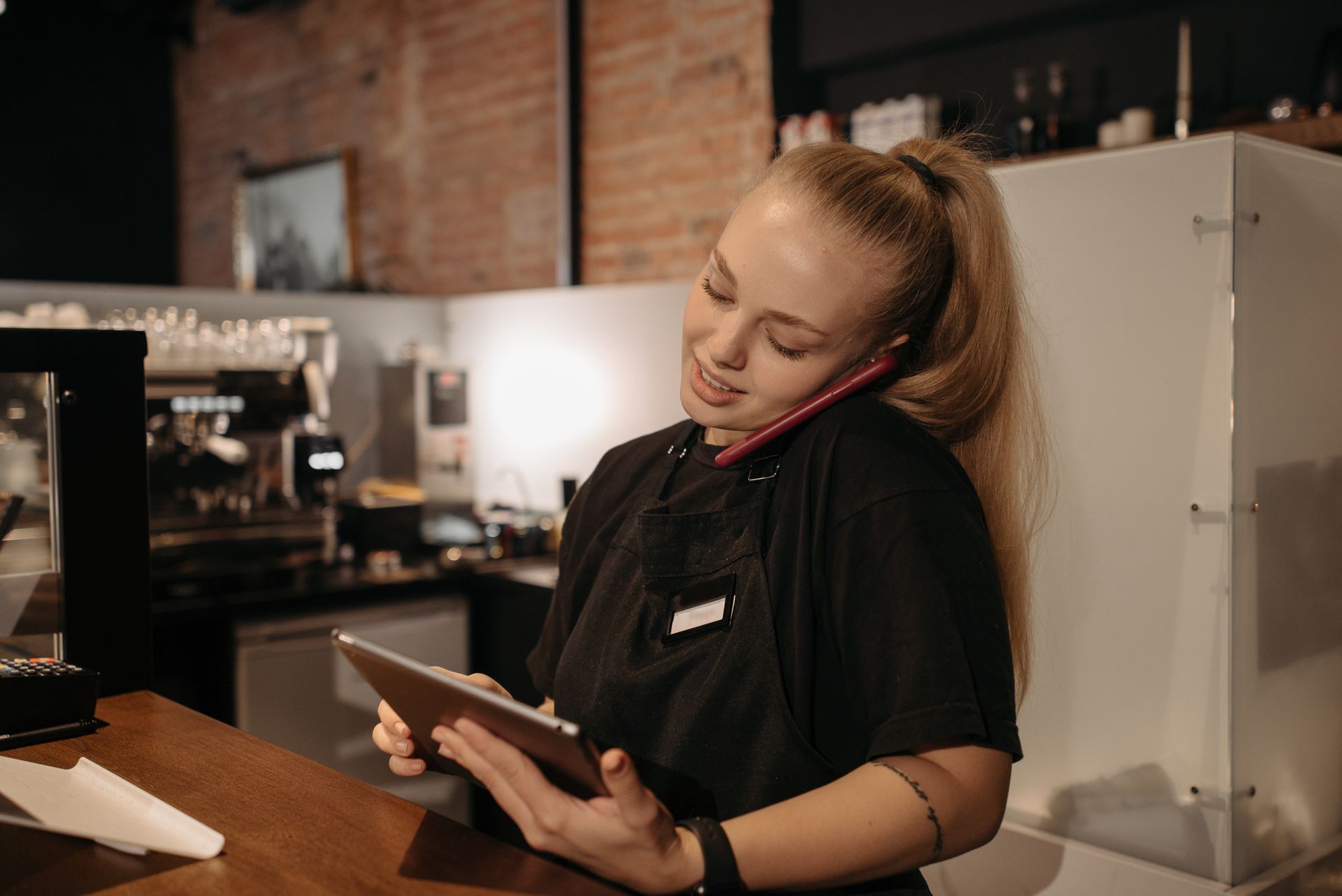 Barista wanita sedang menelepon di ponsel dan memegang tablet di meja bar kafe