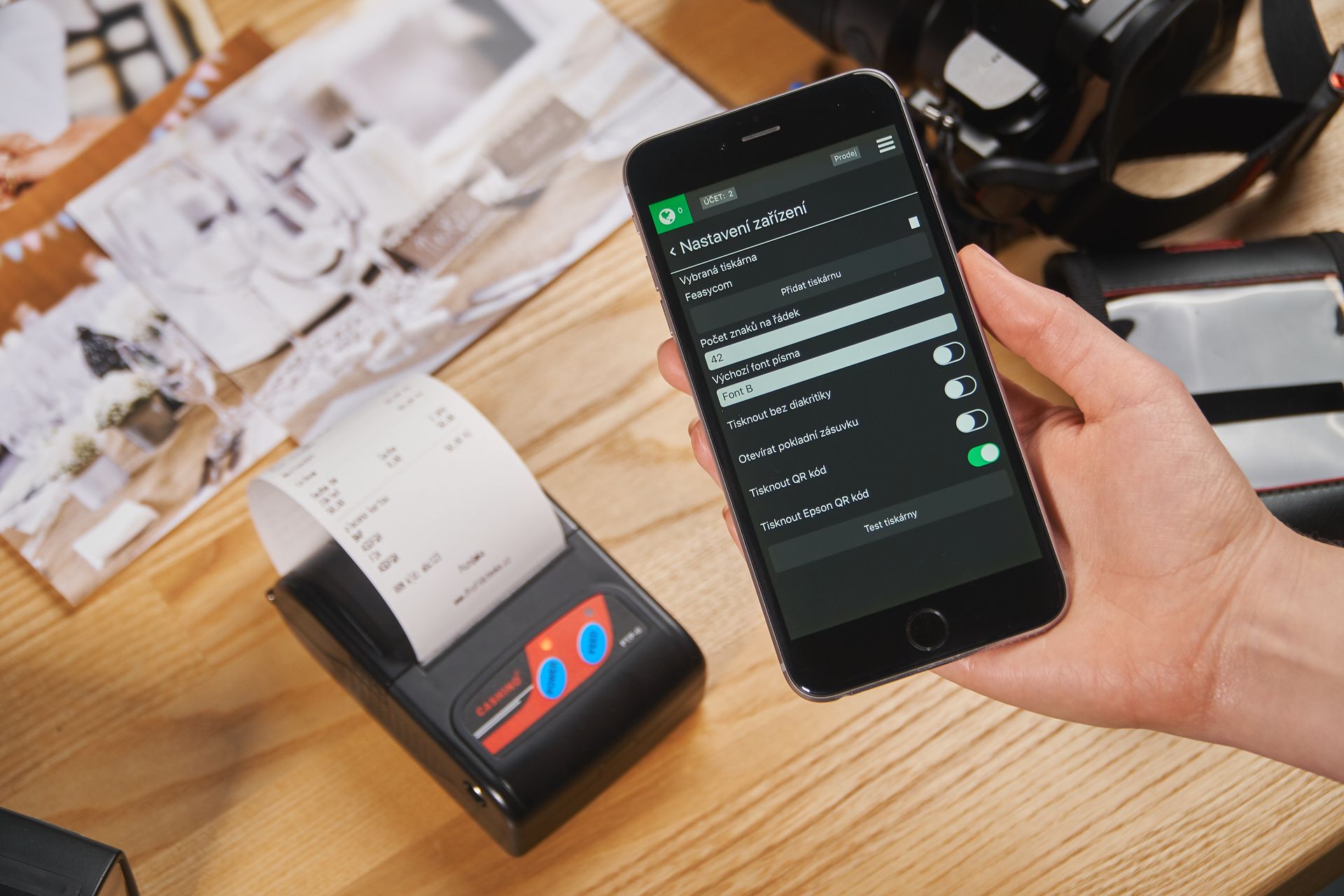 Pengaturan-Aplikasi-Mobile-dan-Printer-Thermal-Bluetooth-Kasir Seseorang memegang smartphone dengan aplikasi kasir dan printer thermal kecil mencetak struk