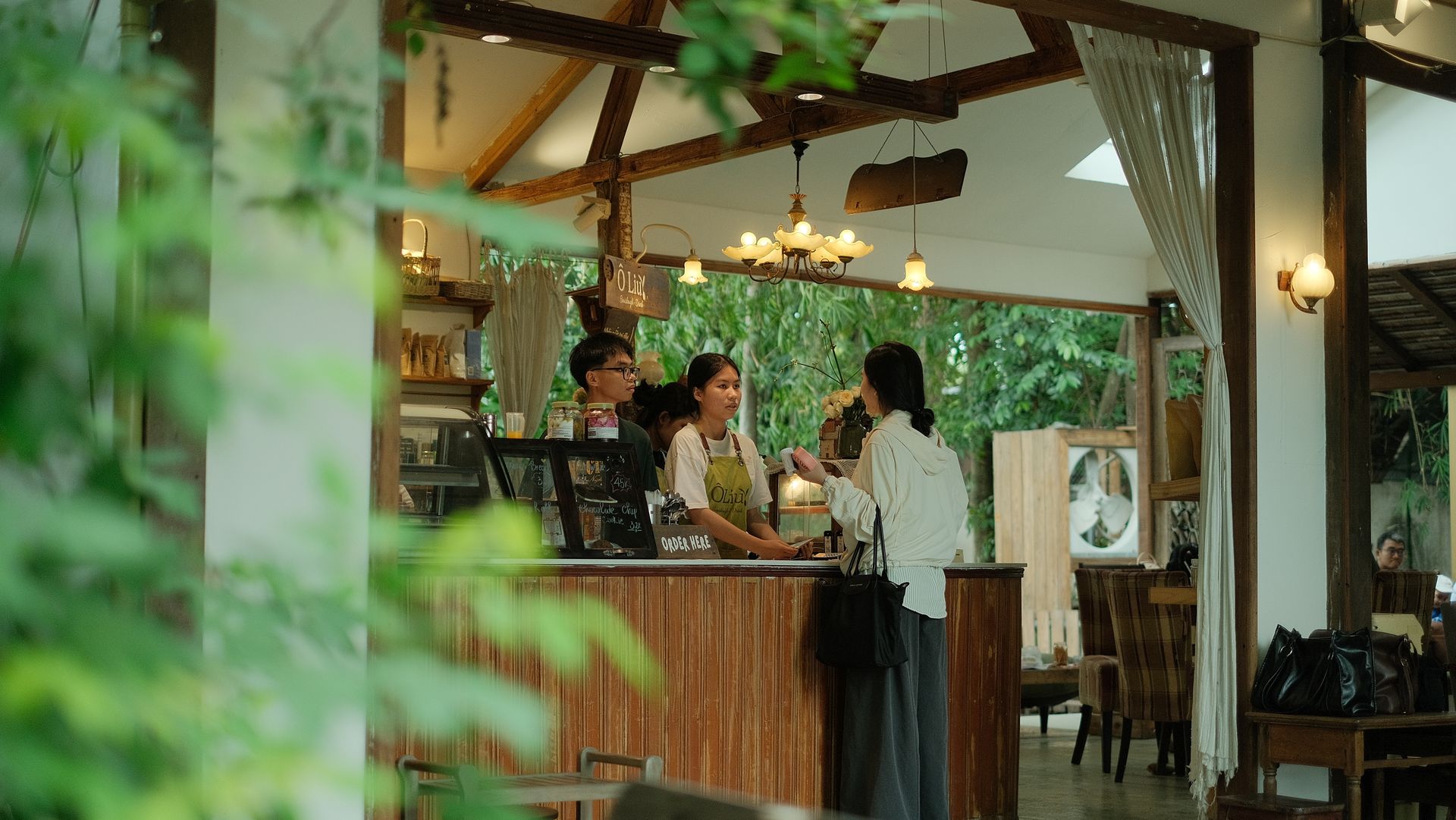 Pelayanan Kasir dan Pemesanan Pelanggan di Kafe Outdoor Pelanggan wanita memesan makanan di counter kayu kafe bernuansa alam yang dilayani oleh dua barista berapron