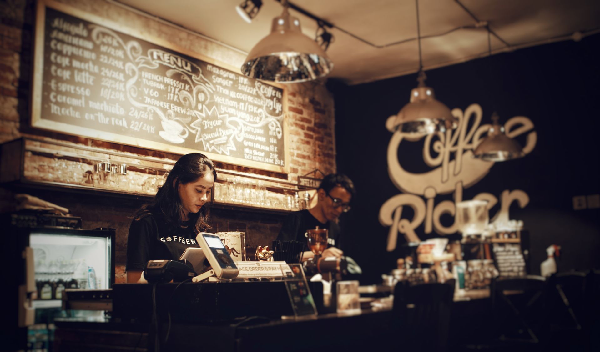 Barista wanita bekerja di balik meja kasir dengan latar belakang papan menu tulis kapur dan tulisan Coffee Rider