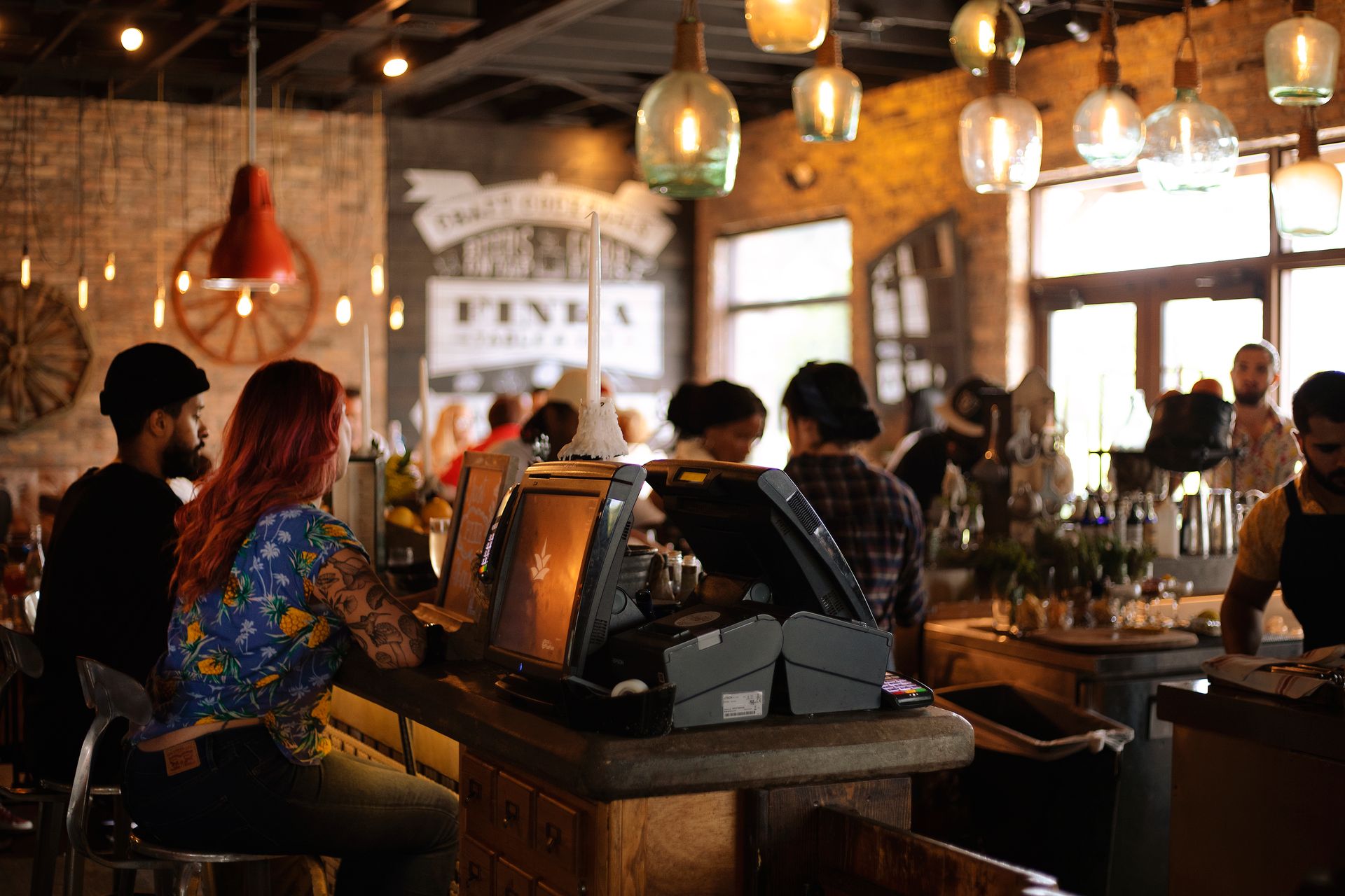 Suasana-Ramai-di-Bar-atau-Kafe-dengan-Pencahayaan-Hangat Pengunjung duduk di bar dan konter kasir dengan lampu gantung dekoratif yang estetik di dalam kafe