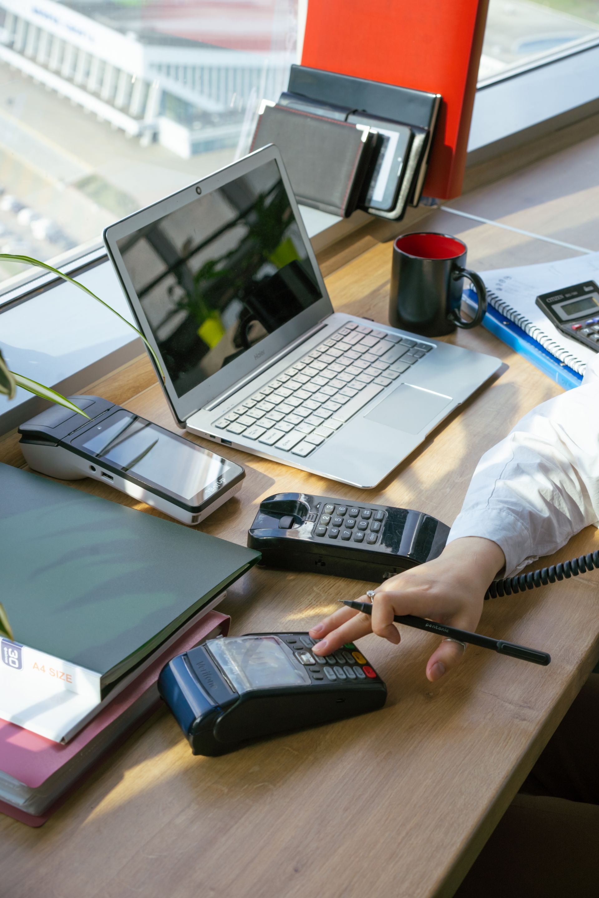 Aktivitas-Pekerja-Kantor-Menggunakan-Perangkat-Kasir-dan-Laptop-di-Meja Seseorang bekerja di meja kantor di sebelah laptop, mesin EDC, dan buku catatan dekat jendela