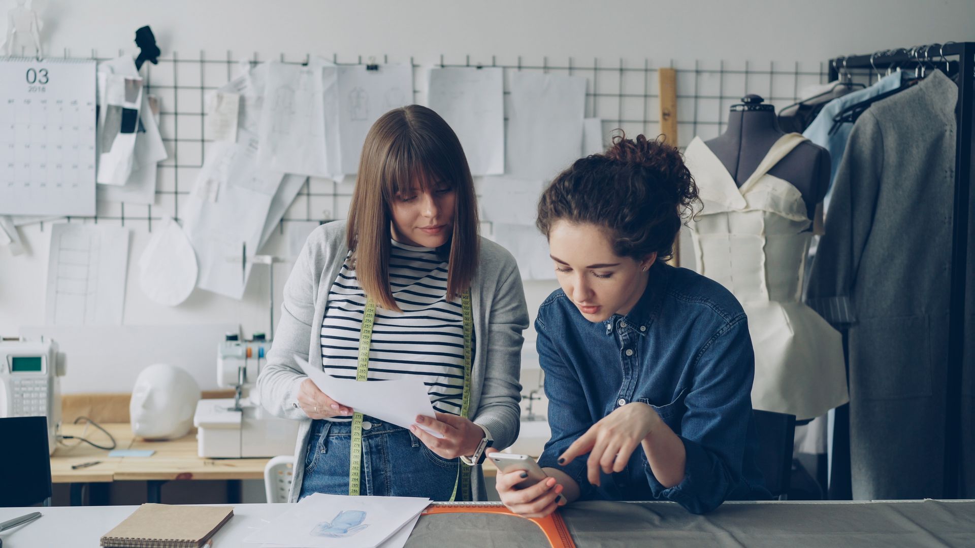 Dua-Perancang-Busana-Berdiskusi-Mengenai-Desain-di-Studio-Konveksi Dua wanita perancang busana bekerja di studio dengan papan sketsa, manekin, dan melihat ponsel pintar