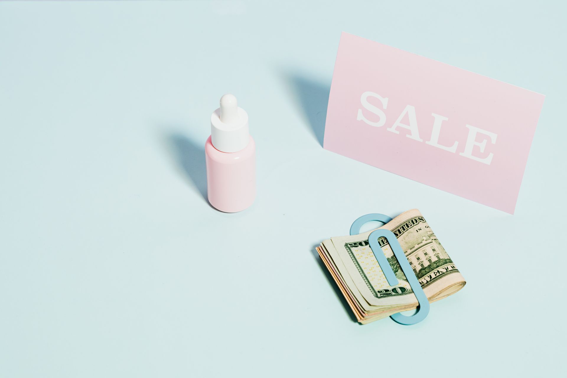 Konsep-Diskon-Produk-Kecantikan-dan-Uang-Tunai-di-Atas-Meja Botol pipet skincare merah muda, kartu bertuliskan SALE, dan segepok uang dolar dengan penjepit kertas di atas latar biru