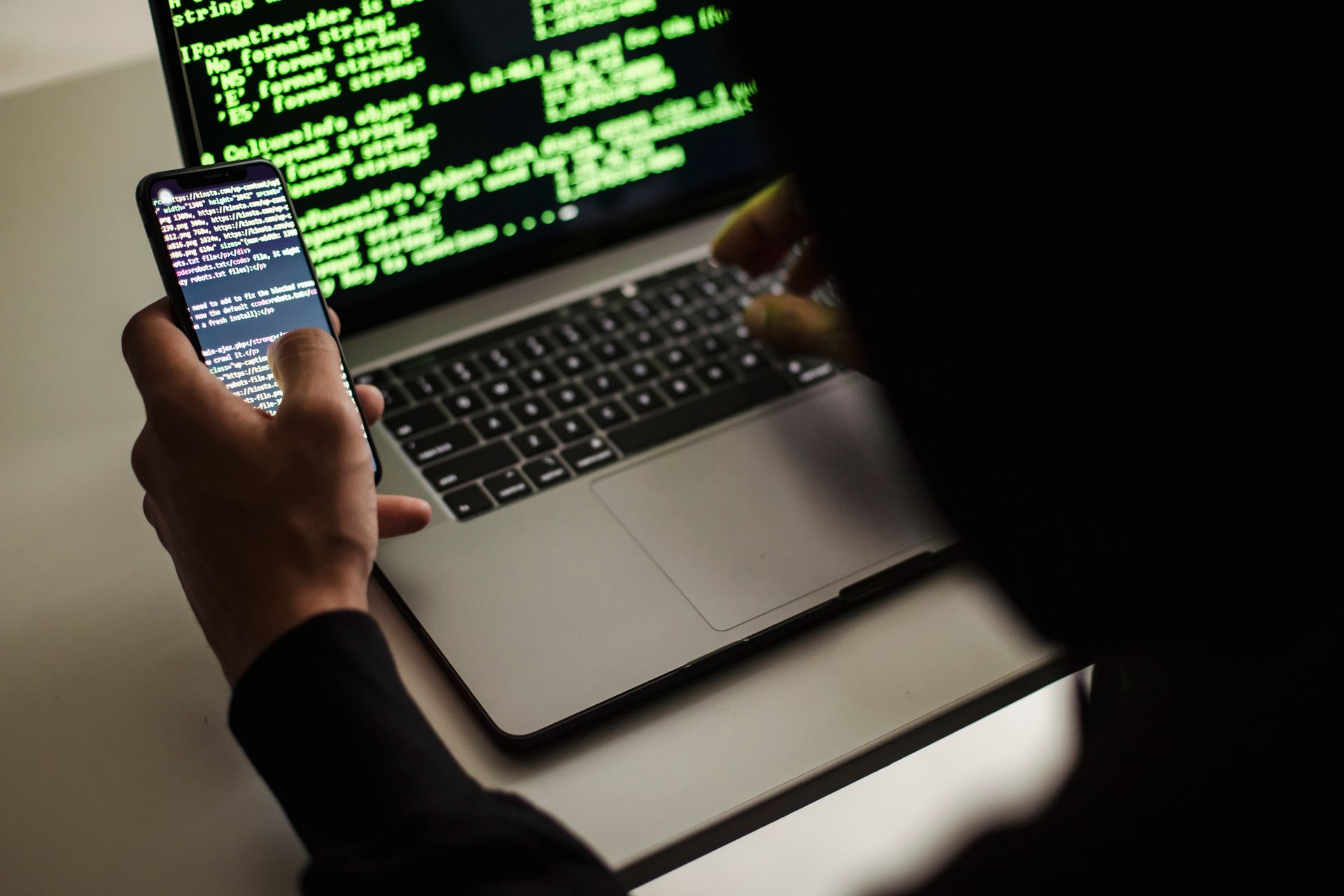 keamanan-data Seseorang memegang ponsel pintar sambil mengetik di laptop yang menampilkan deretan kode pemrograman berwarna hijau.