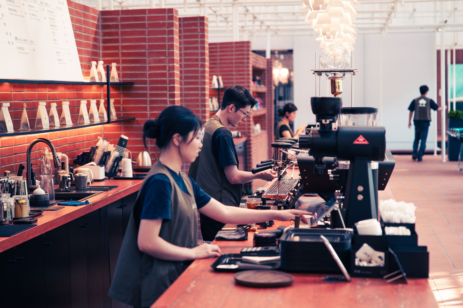 Suasana-Sibuk-Barista-Bekerja-di-Kedai-Kopi-Modern Beberapa barista sedang melayani dan meracik kopi di balik meja bar dengan latar dinding bata merah
