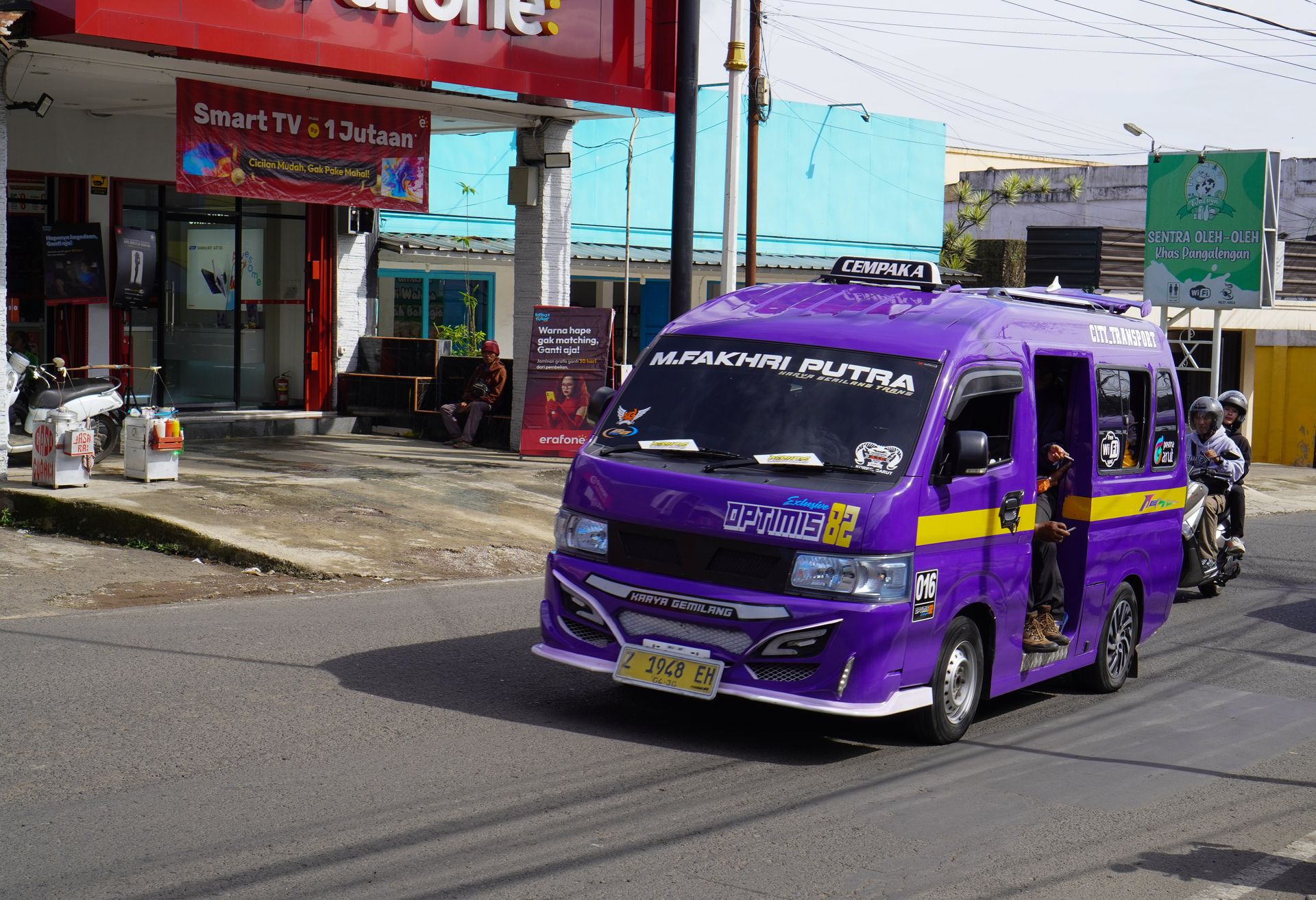 Sebuah mobil angkutan umum angkot berwarna ungu dengan modifikasi dan tulisan M. FAKHRAI PUTRA melintas di jalan raya