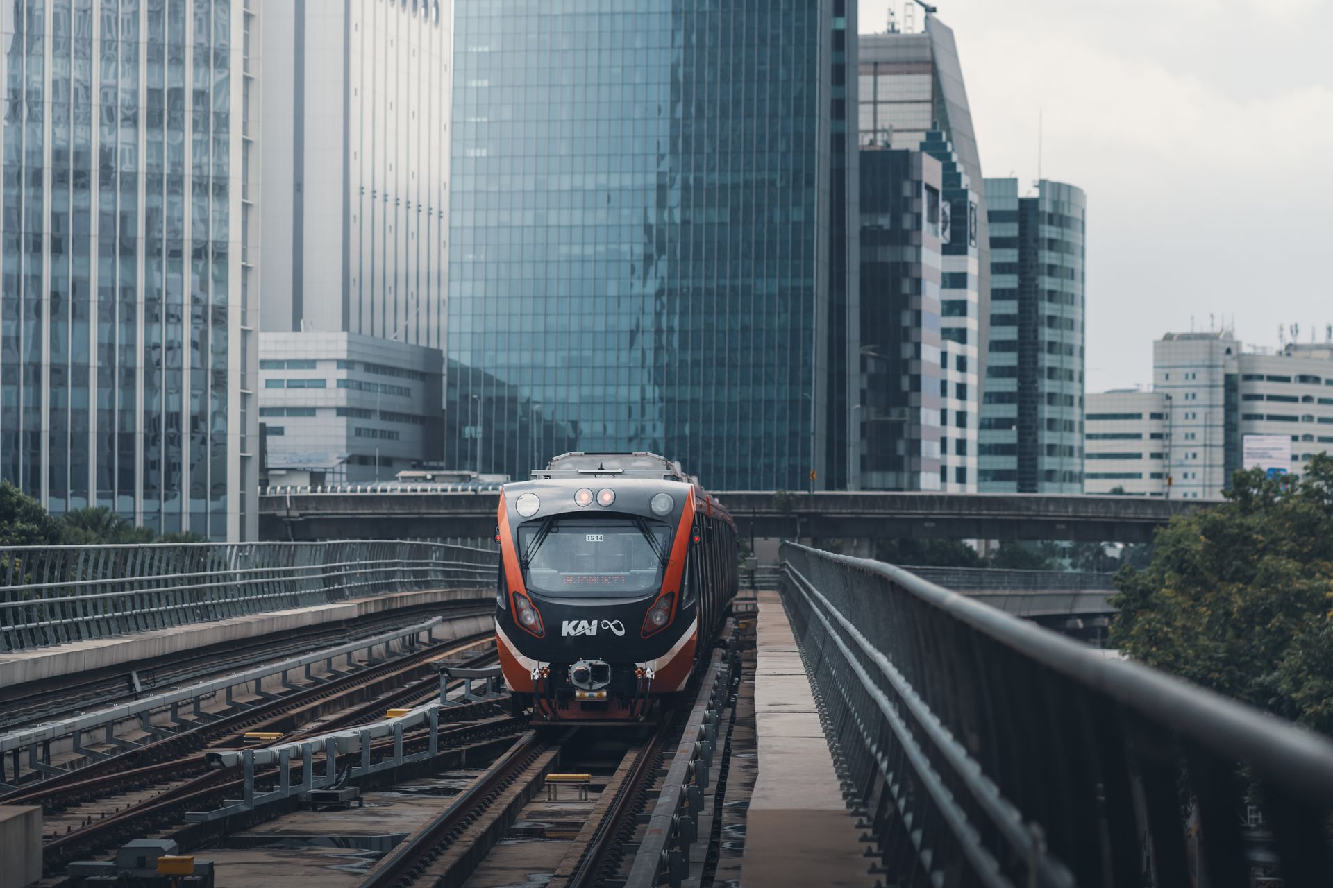 Kereta-Api-KAI-Berjalan-di-Jalur-Elevasi-Perkotaan Sebuah kereta api berlogo KAI melintas di jalur rel layang dengan latar belakang gedung-gedung pencakar langit perkotaan