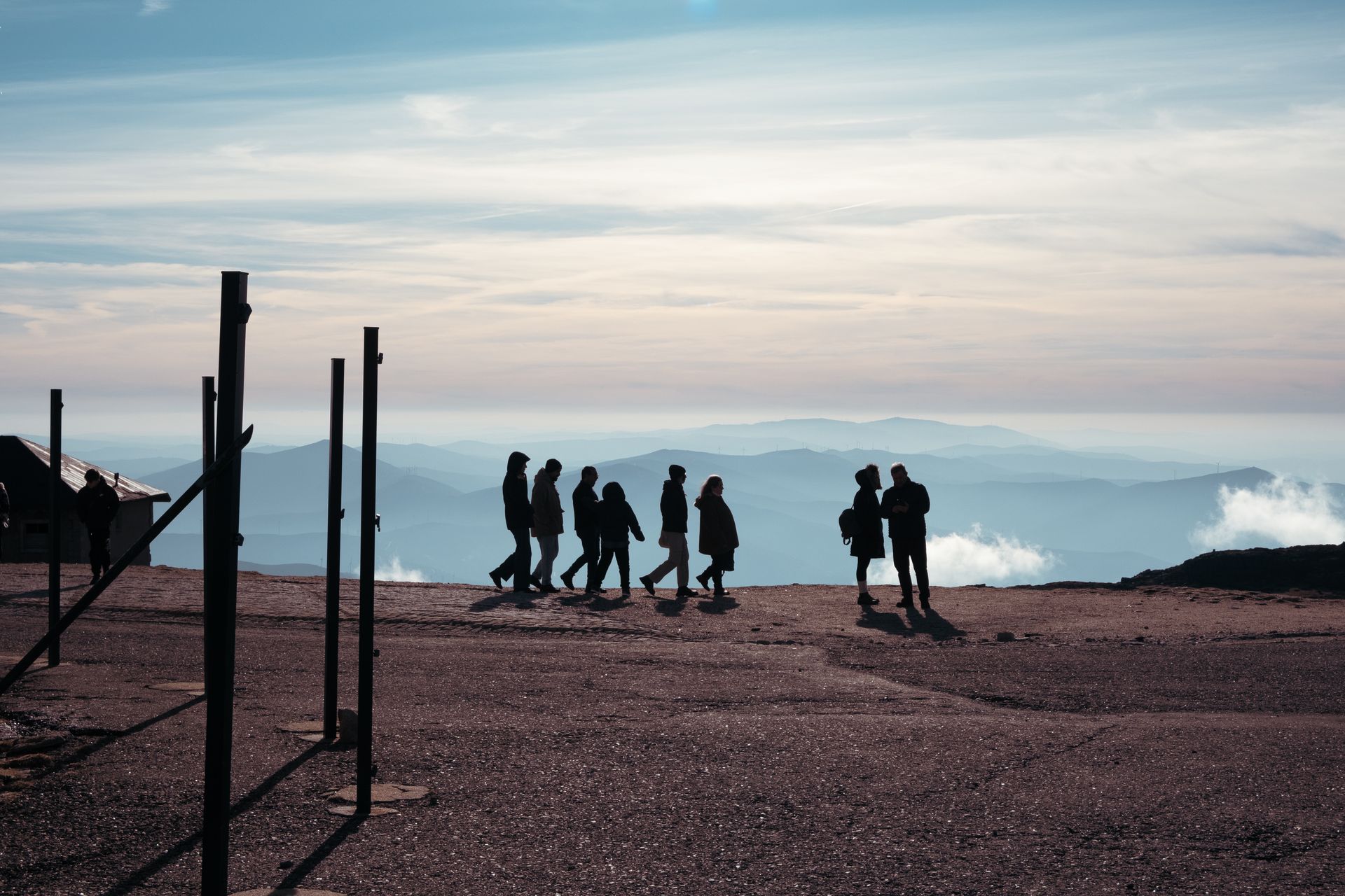 Siluet sekelompok pendaki dan wisatawan sedang berjalan di puncak gunung dengan pemandangan perbukitan dan awan.