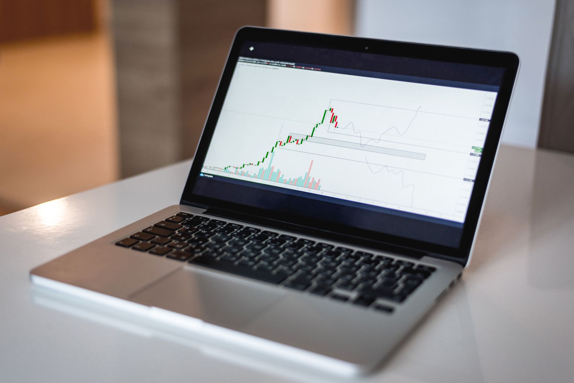 Laptop-Menampilkan-Grafik-Trading-dan-Analisis-Pasar-Saham Sebuah laptop perak terbuka di atas meja putih dengan layar menampilkan grafik trading dan pergerakan harga saham