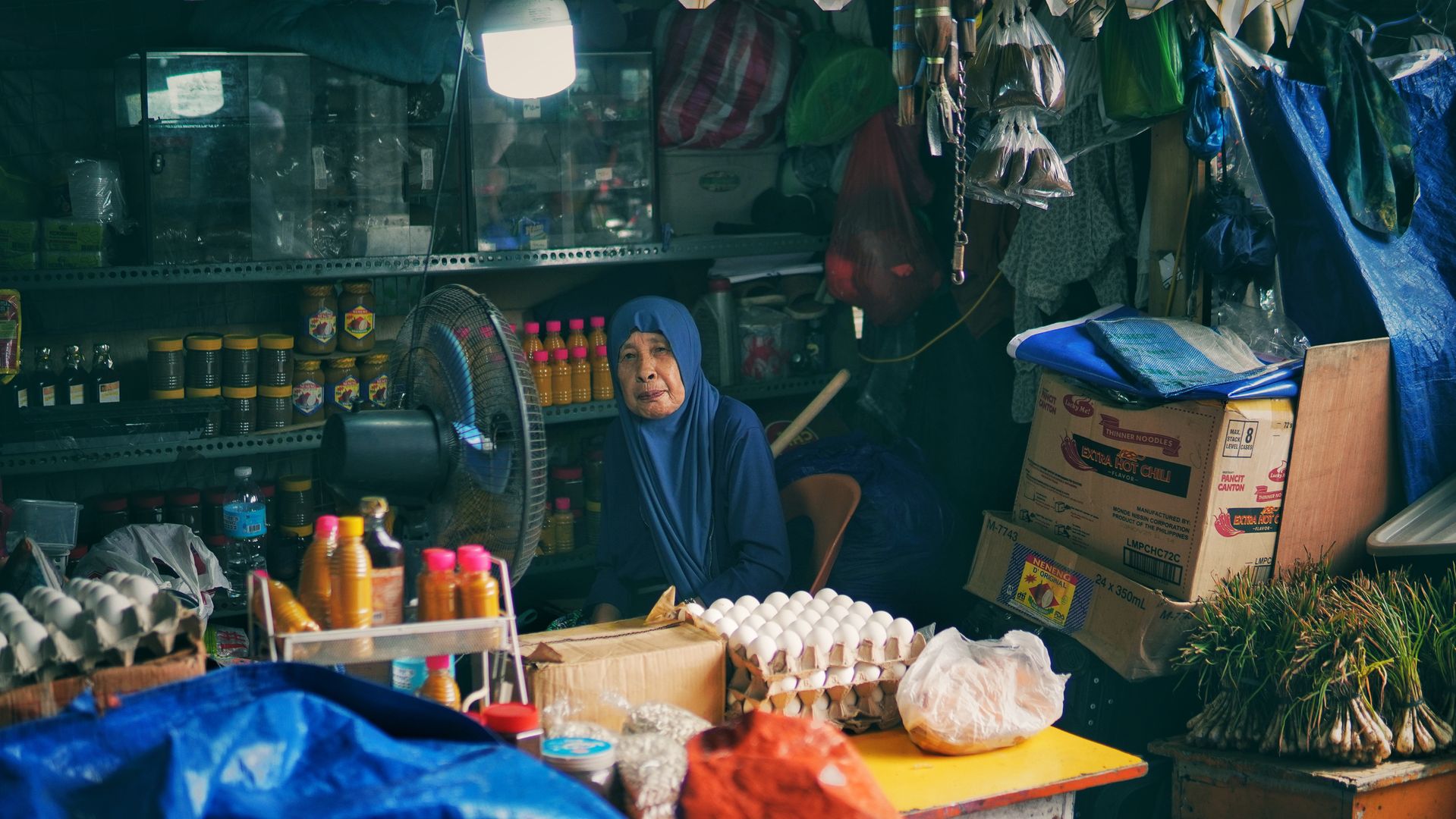 Seorang-Ibu-Menjaga-Warung-Kelontong-Tradisional Seorang wanita paruh baya mengenakan kerudung biru duduk menjaga warung kelontong tradisional yang penuh dengan barang dagangan