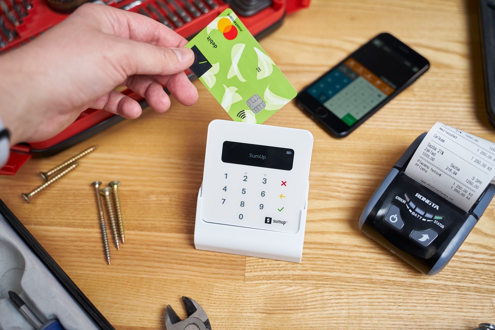 Pembayaran Contactless Kartu Debit di Perangkat Mobile POS Portabel Tangan menempelkan kartu debit Mastercard hijau di atas perangkat pembaca kartu portabel putih lengkap dengan printer thermal dan peralatan teknisi di atas meja kayu