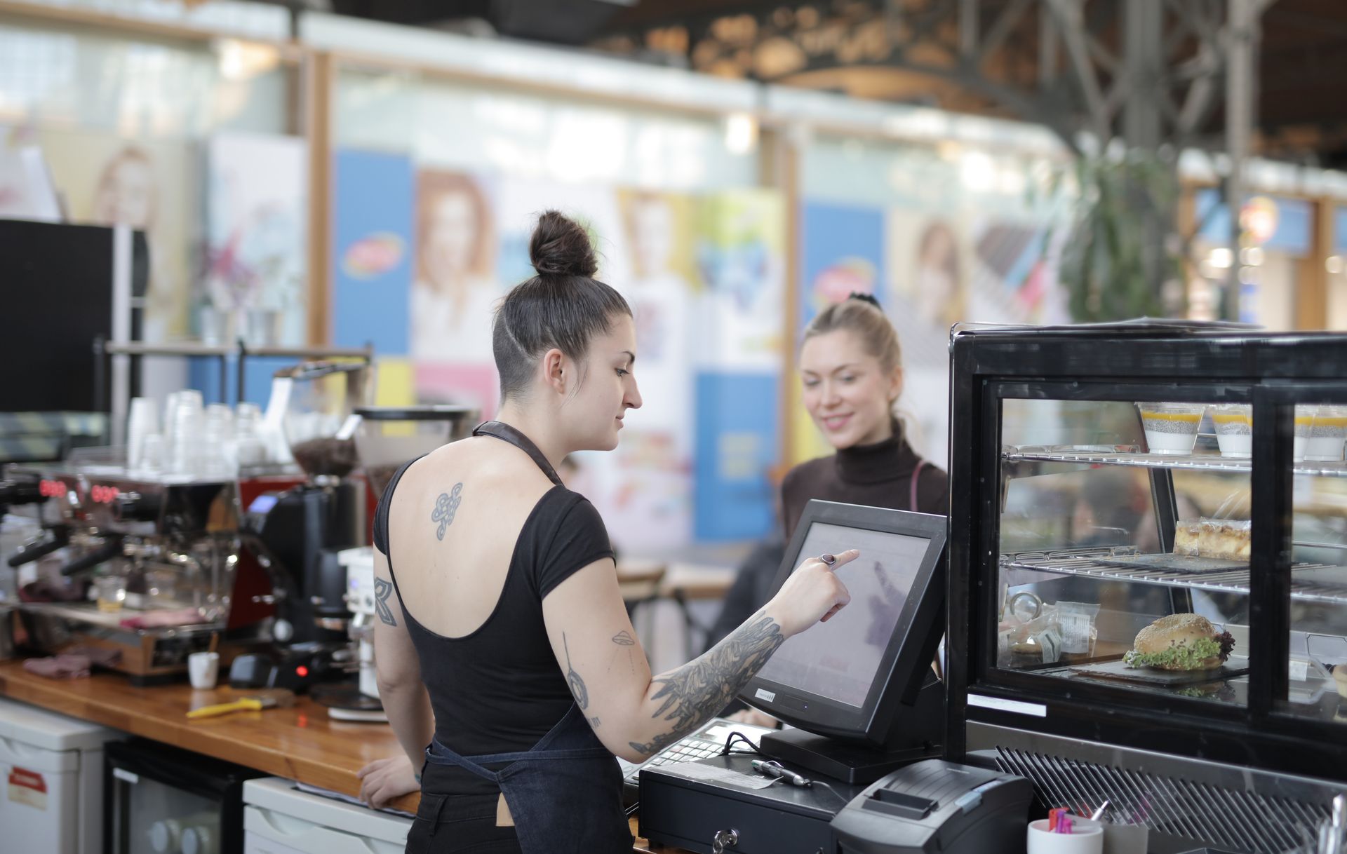 Kasir-Wanita-Mengoperasikan-Layar-Sentuh-POS-di-Kafe Seorang wanita berambut sanggul bertato mengoperasikan layar sentuh mesin kasir POS di balik meja bar kafe