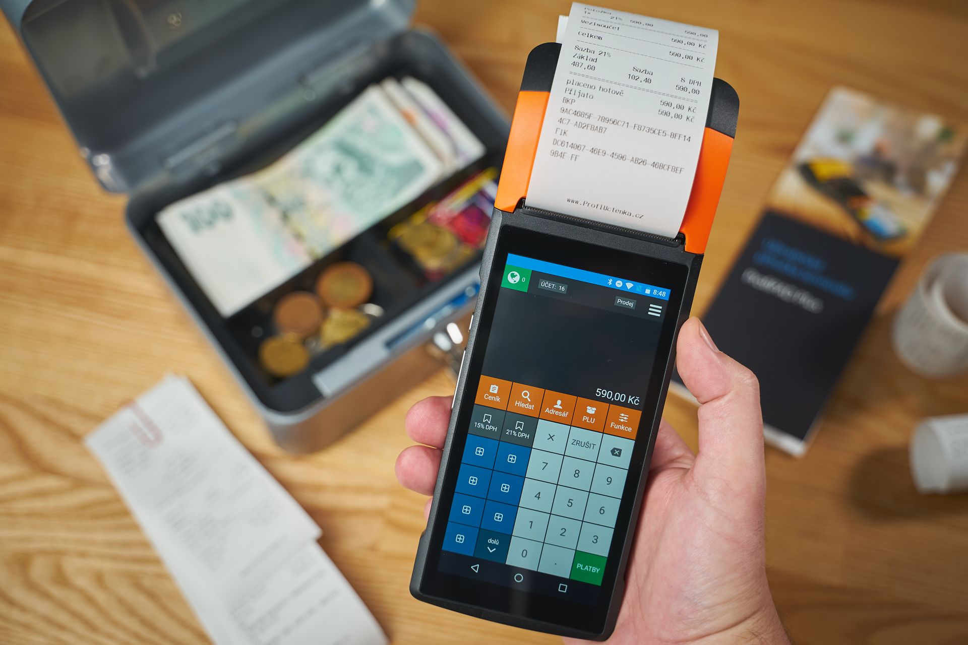 Mesin-EDC-Mobile-POS-dan-Kotak-Penyimpanan-Uang-Tunai Seseorang memegang mesin EDC mobile POS yang mencetak struk belanja, di sebelah kotak uang kasir yang terbuka berisi uang tunai