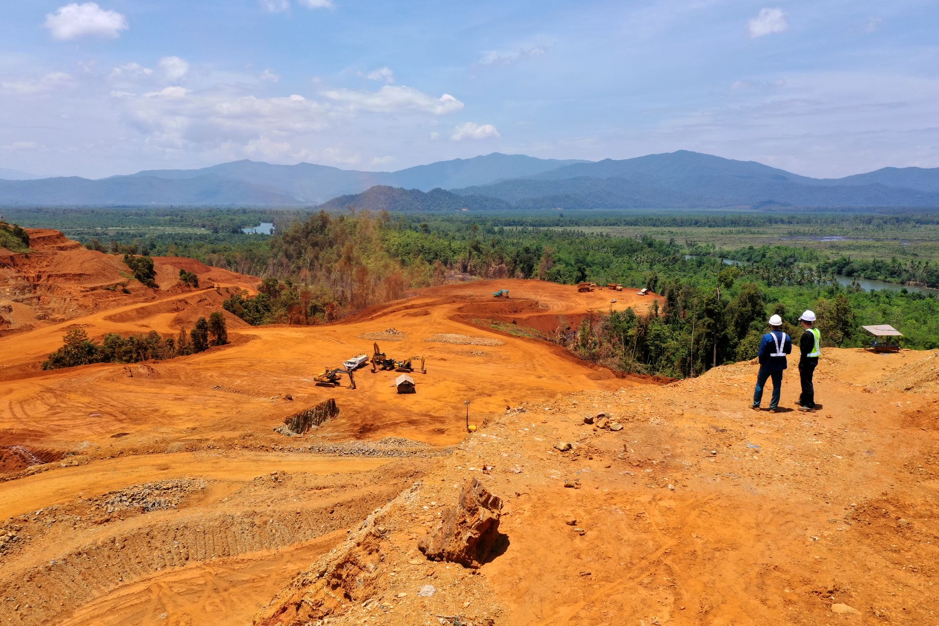 Inspeksi-Lokasi-Pertambangan-Terbuka-di-Area-Pegunungan Dua orang insinyur mengenakan helm pengaman berdiri di atas bukit mengamati area tambang terbuka berTanah merah dengan latar pegunungan