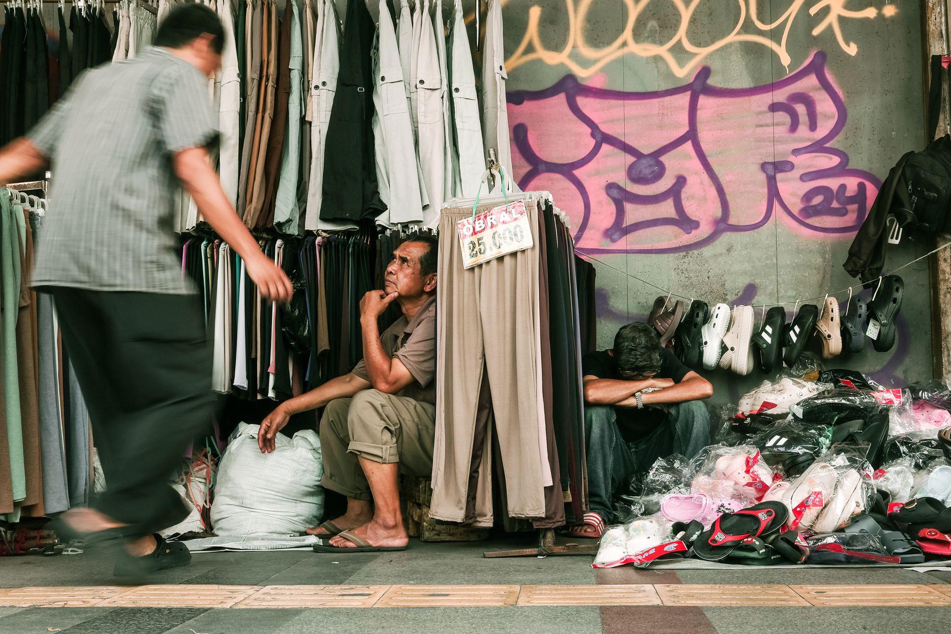 Suasana-Kios-Pakaian-dan-Alas-Kaki-di-Pinggir-Jalan Seorang penjaga duduk termenung di kios pakaian kaki lima berlatar dinding bergambar grafiti dengan tumpukan alas kaki dan baju gantung