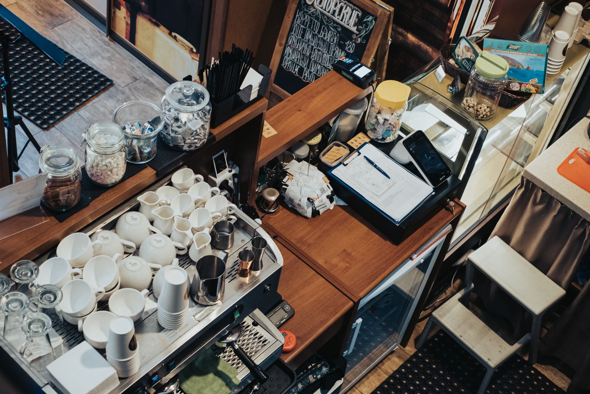 Foto dari atas (high-angle) memperlihatkan area bar kafe yang berisi mesin kopi, tumpukan cangkir putih, toples, dan meja kasir