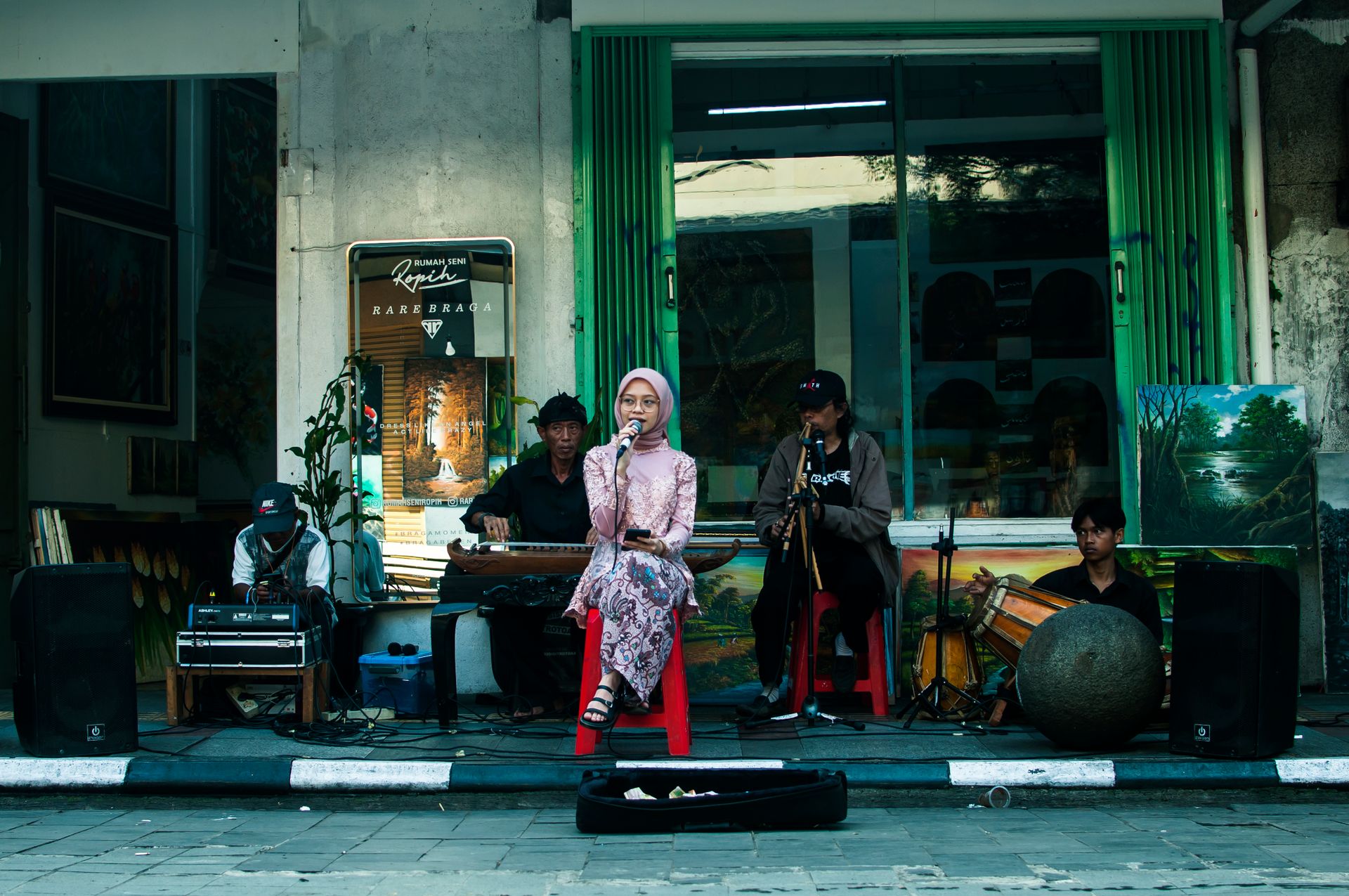 Grup-Musik-Jalanan-Tampil-di-Depan-Ruko Seorang wanita bernyanyi di depan sekelompok pemusik jalanan tradisional yang memainkan alat musik di trotoar depan ruko