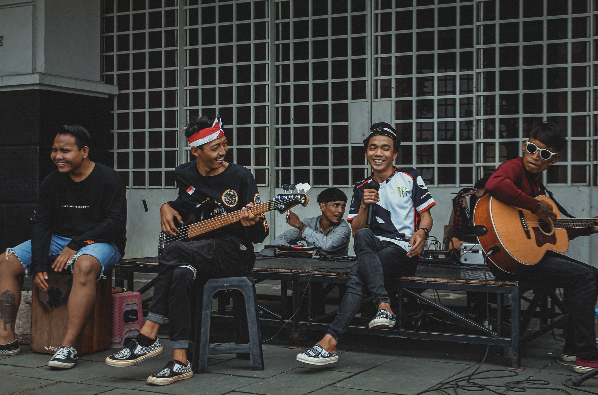 Grup-Musik-Jalanan-Pria-Tampil-Bersama-di-Trotoar Sekelompok pemuda pemusik jalanan bermain gitar, bass, dan perkusi di atas panggung kecil di trotoar