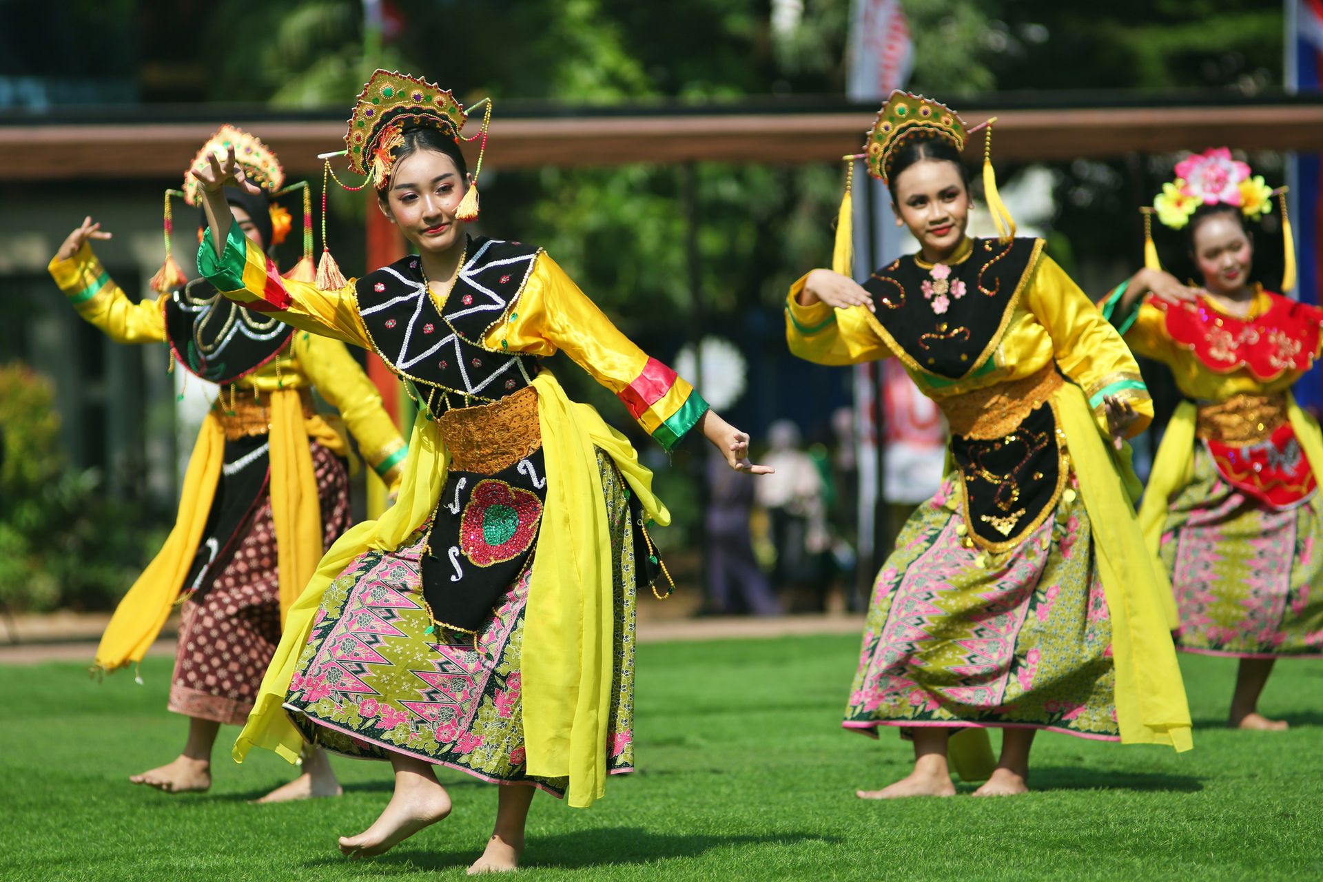 Penari-Tradisional-Wanita-Tampil-di-Area-Terbuka Sekelompok penari wanita mengenakan pakaian tradisional berwarna kuning dan kain corak menari di atas lapangan rumput hijau