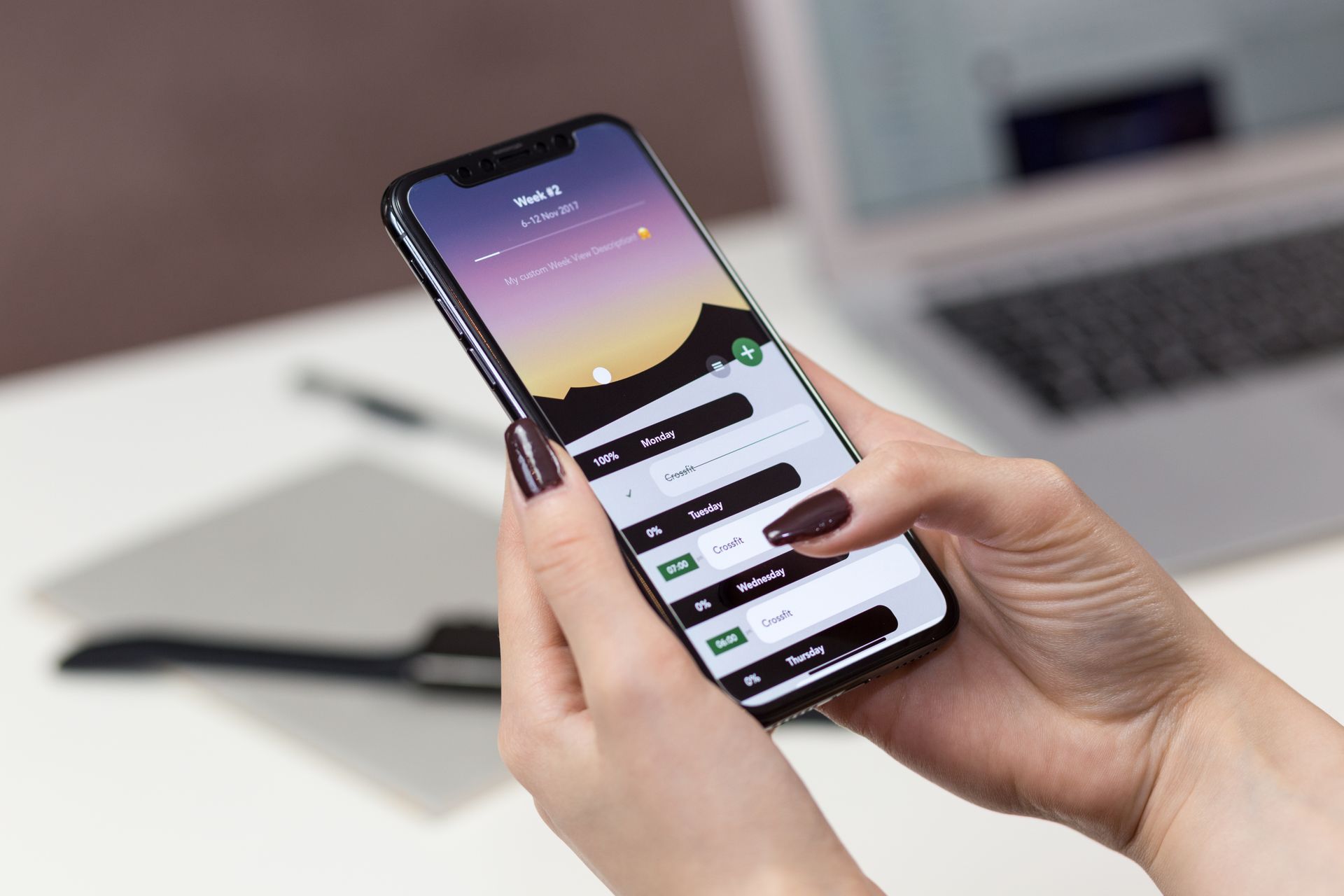 Seseorang-Mengoperasikan-Aplikasi-Perencanaan-di-Ponsel Seseorang memegang smartphone menampilkan antarmuka aplikasi jadwal di atas meja dekat laptop terbuka