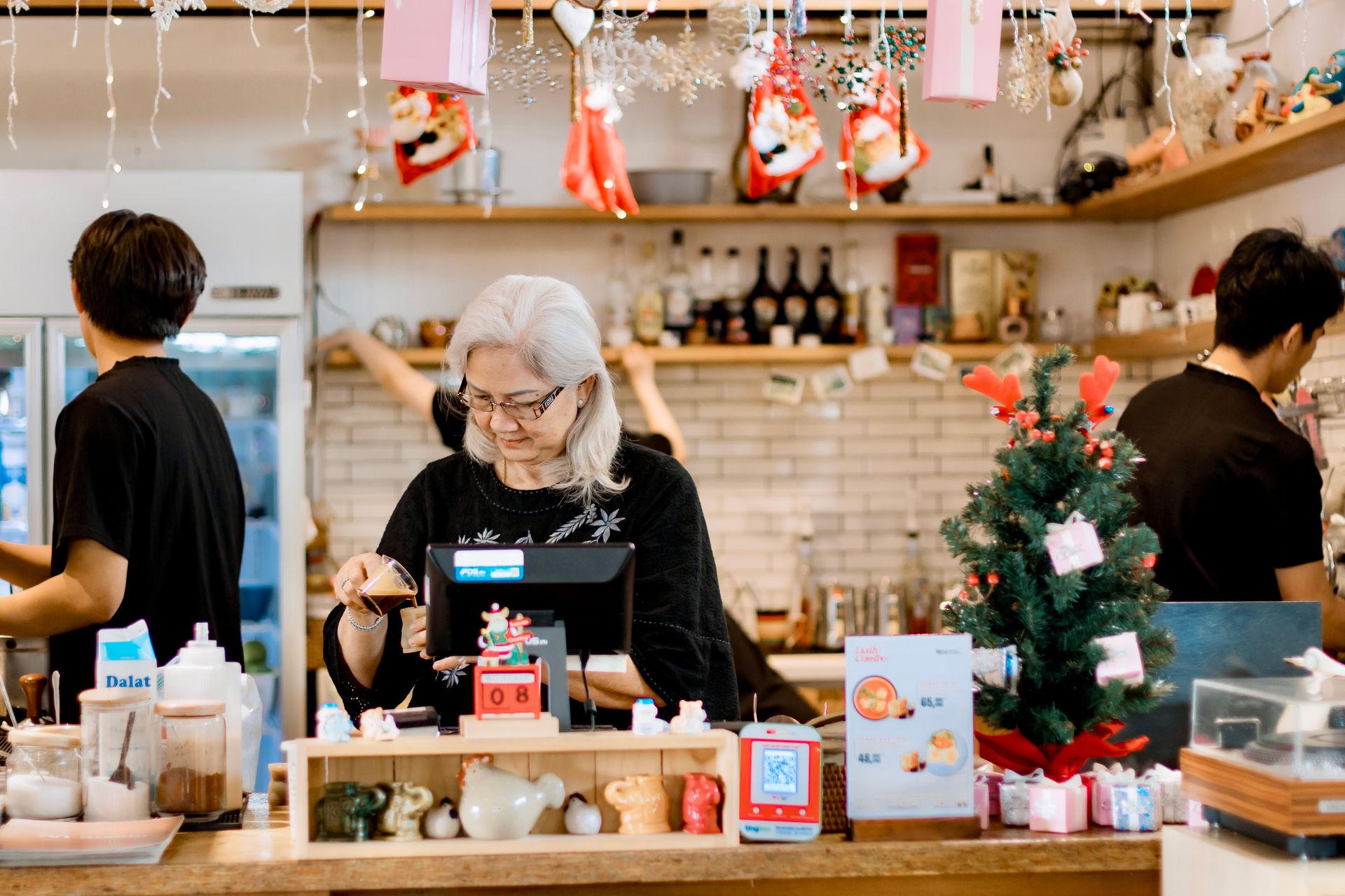 Suasana-Kasir-dan-Barista-di-Kedai-Nuansa-Natal Seorang wanita paruh baya berambut putih melayani di balik meja kasir kafe dengan dekorasi pohon natal kecil dan lampu gantung