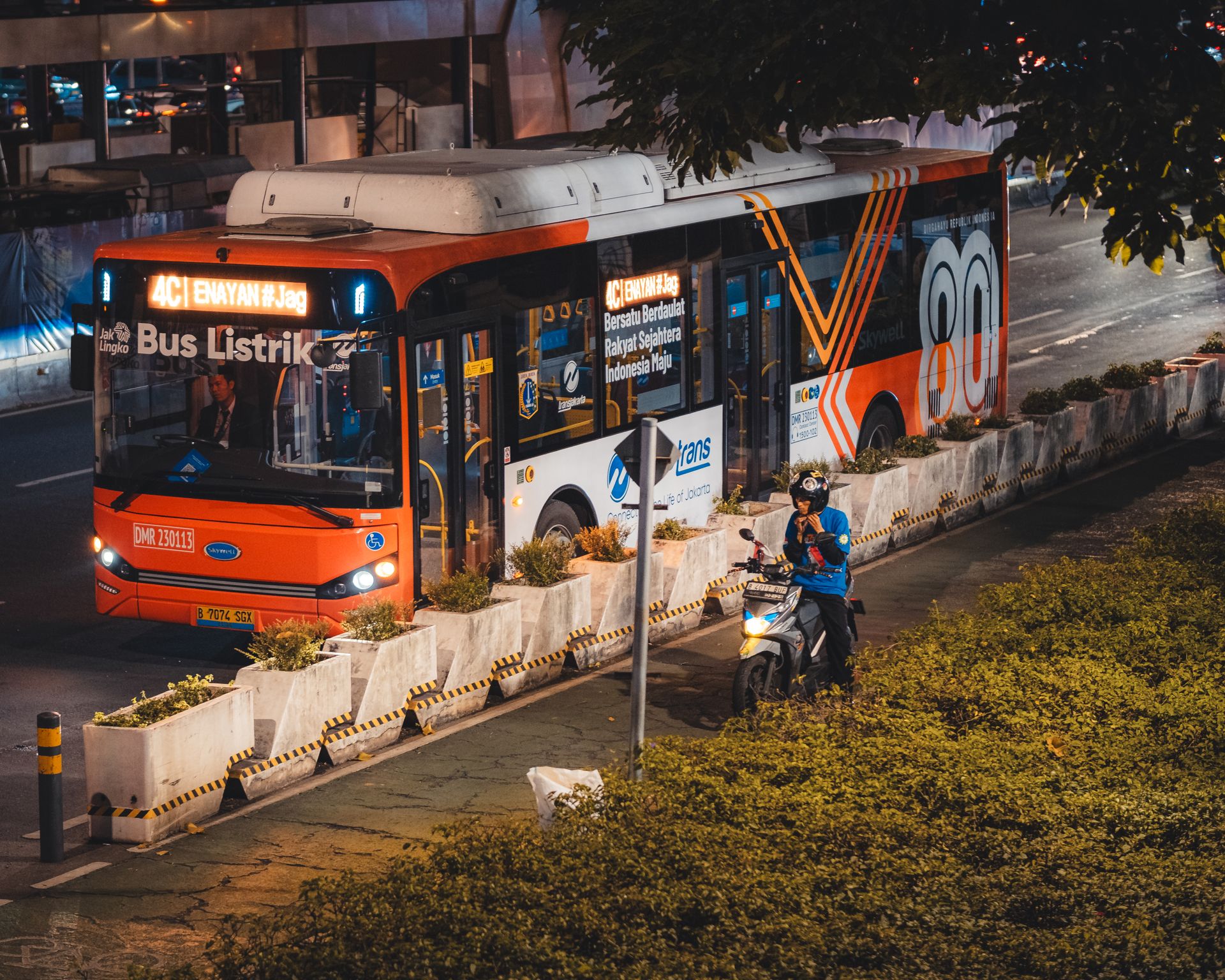 Armada Bus Listrik Transjakarta dalam Layanan Transportasi Publik Bus listrik Transjakarta oranye rute 4C Senayan melintas di jalan raya perkotaan pada malam hari dekat pengendara sepeda motor