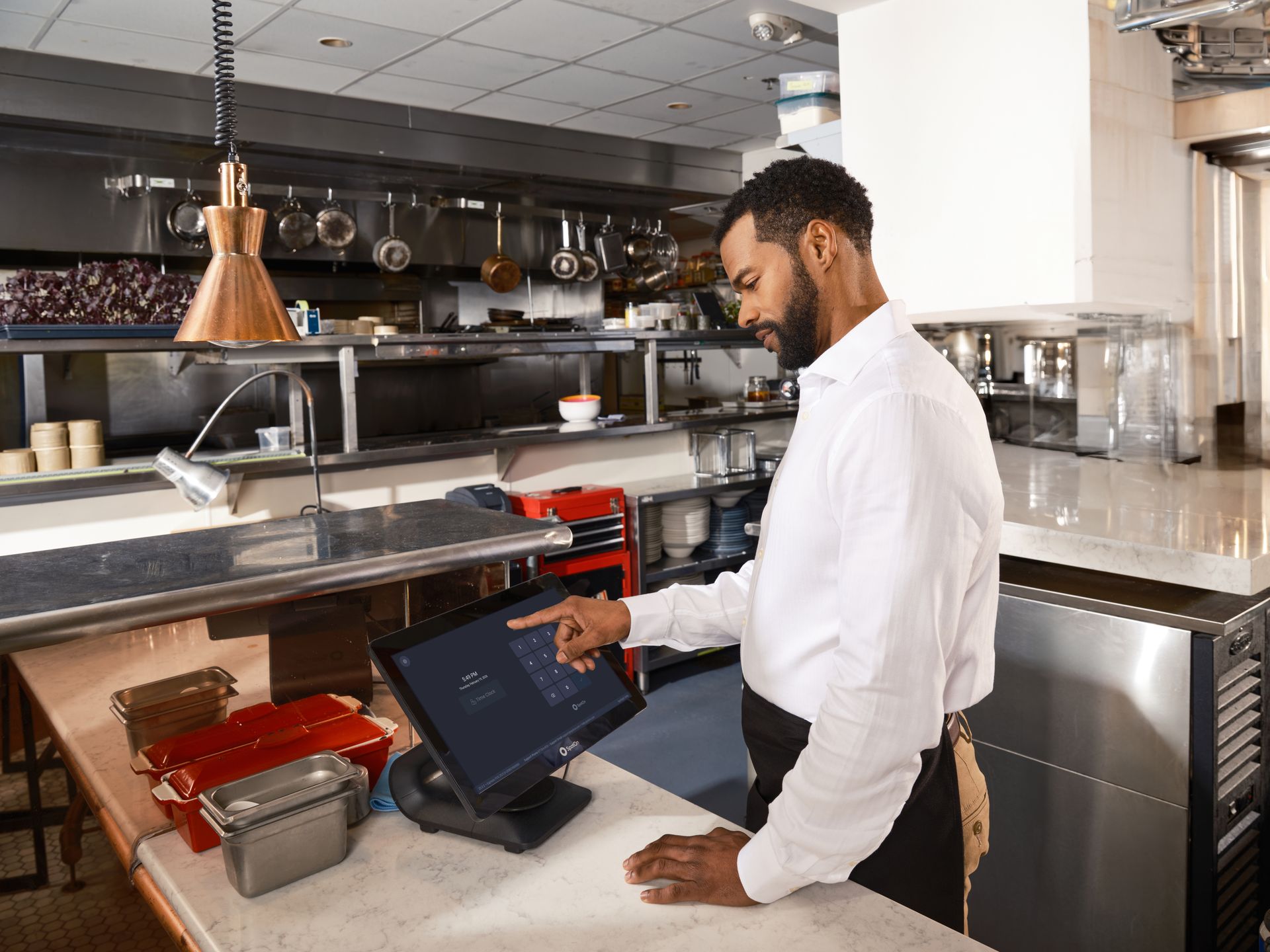 Staf restoran berkemeja putih dan celemek mengoperasikan layar sentuh terminal kasir POS di area dapur profesional