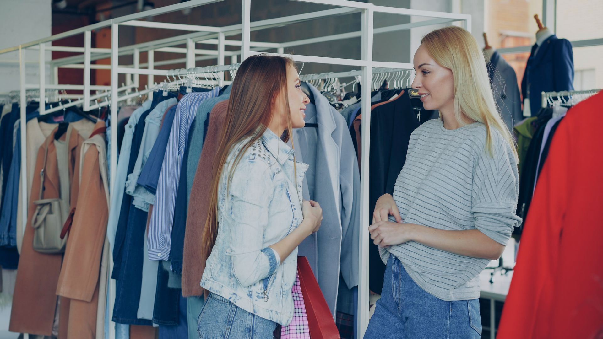 Dua wanita muda berbincang akrab saat berbelanja pakaian di depan deretan gantungan baju butik ritel fashion