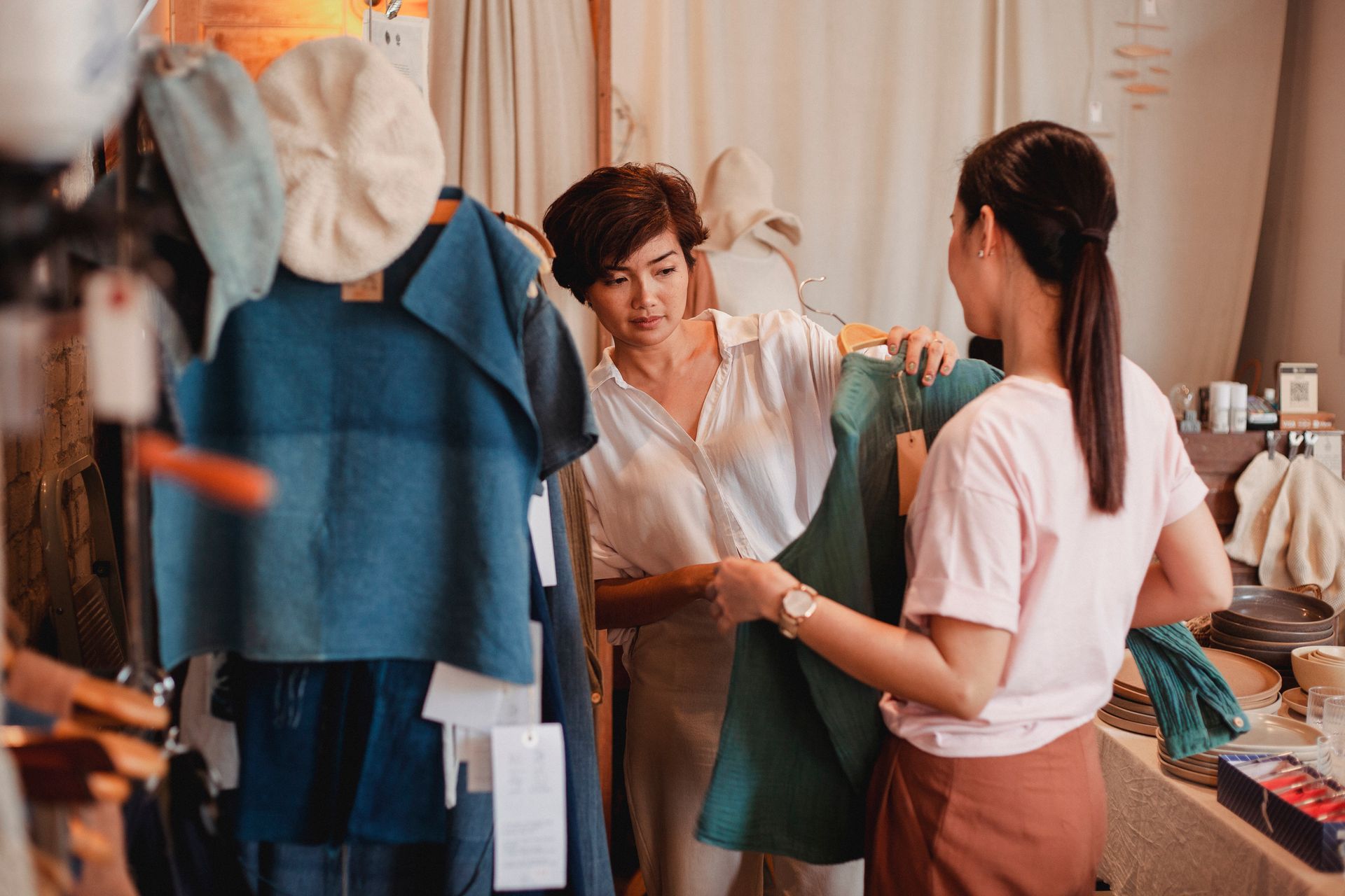 Interaksi Pelanggan Memilih Busana di Butik Ritel Fashion Dua wanita sedang melihat dan memeriksa detail pakaian berwarna hijau pada gantungan baju di butik ritel fashion bernuansa hangat