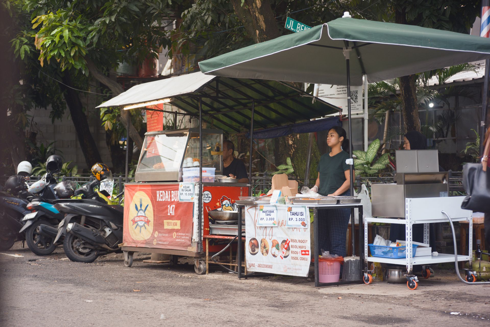 Penjual wanita berdiri di depan lapak gerobak gorengan dan jajanan street food di pinggir jalan.