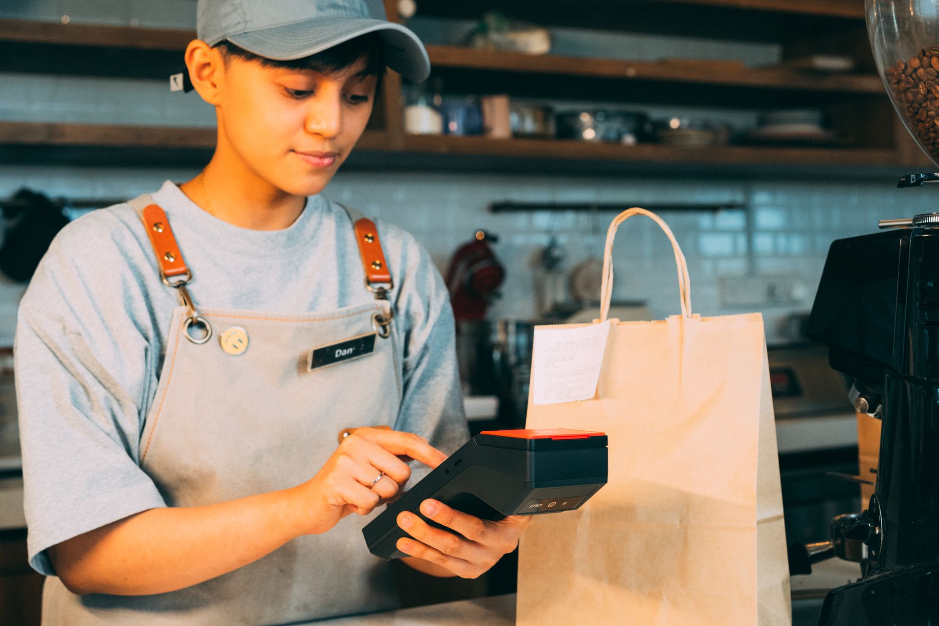 Barista kafe bertopi dan berapron mengoperasikan mesin EDC atau terminal POS portabel di samping kantong kertas belanjaan.