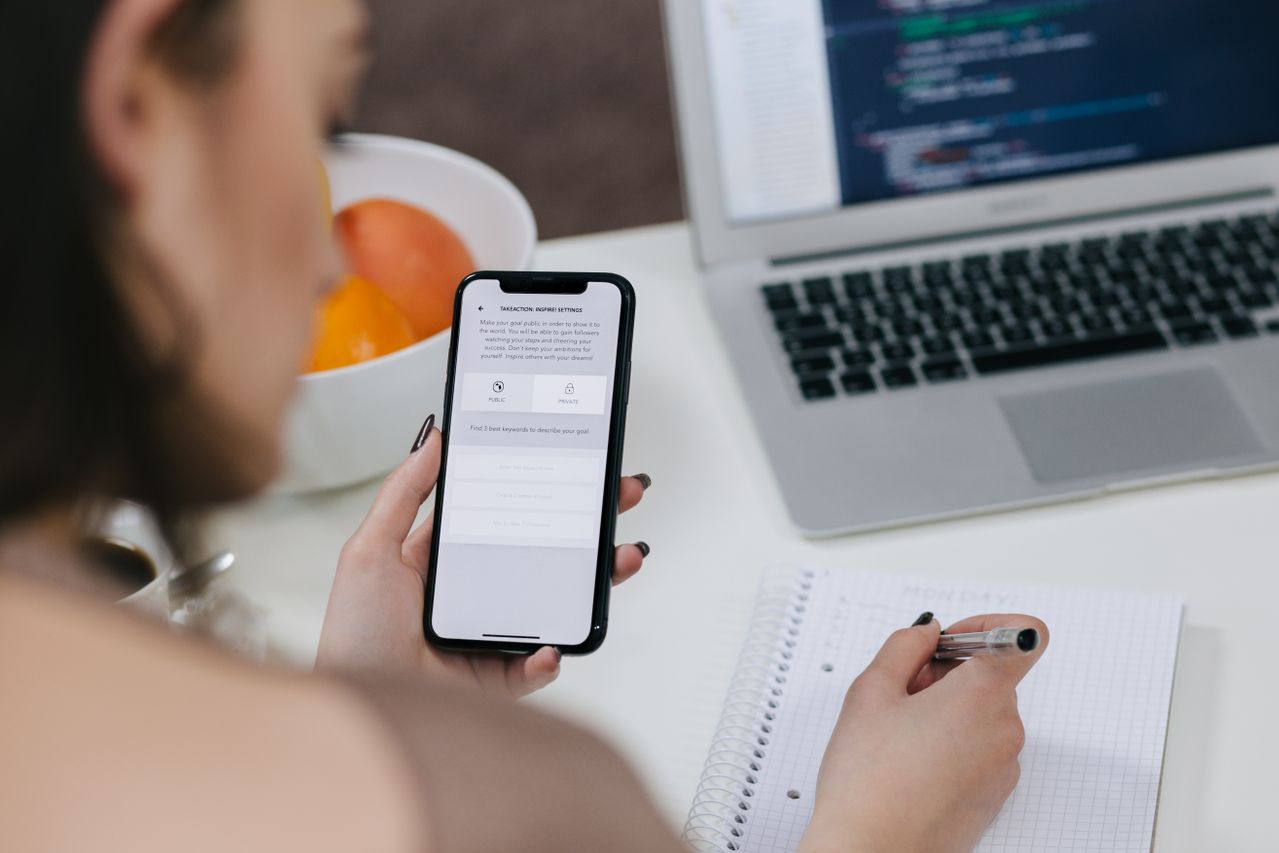 Pengujian dan Monitoring Aplikasi Mobile Kasir (POS) pada Smartphone Tampilan sudut atas bahu seorang wanita mengecek antarmuka aplikasi mobile pada smartphone di dekat buku catatan dan laptop