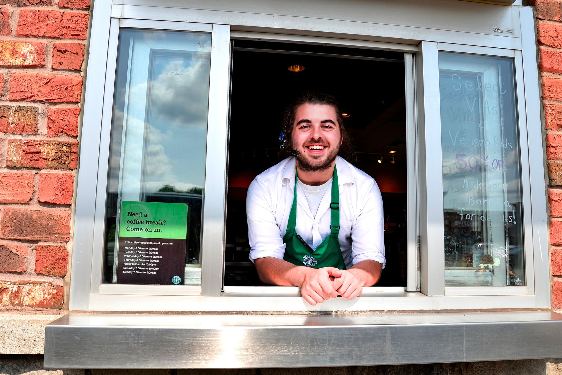Barista pria berapron hijau tersenyum ramah di balik jendela layanan drive-thru kedai kopi.