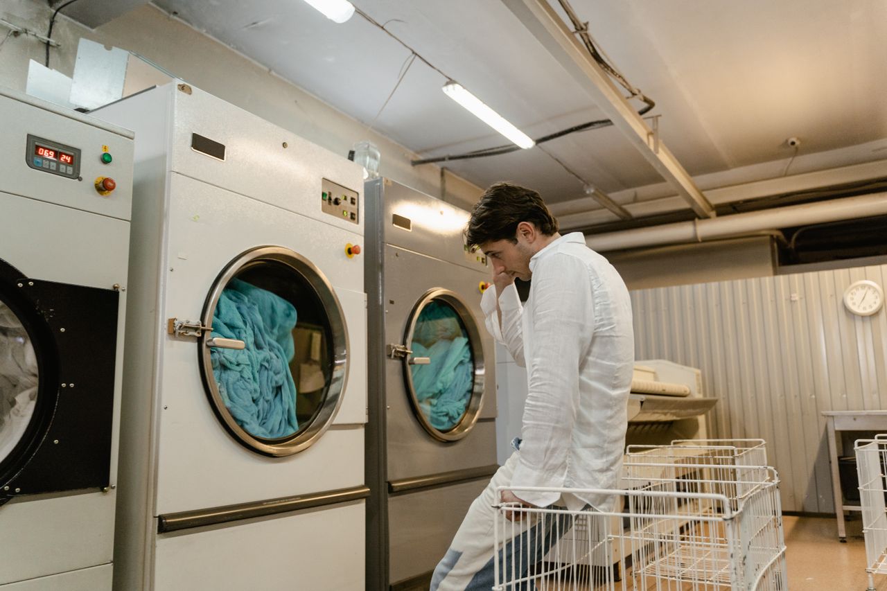 Pengelola memantau operasional mesin cuci industri di tempat usaha menggunakan sistem kasir untuk bisnis laundry Pria duduk bersandar pada keranjang pakaian di dekat deretan mesin cuci industri di ruang operasional bisnis laundry